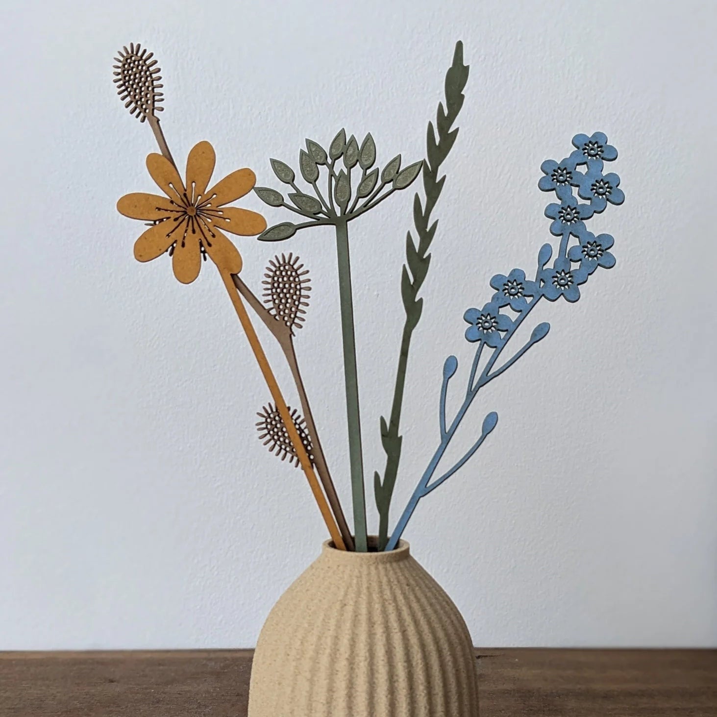 Wooden flower arrangement in a beige vase.  Features orange, blue, and green laser-cut flowers.