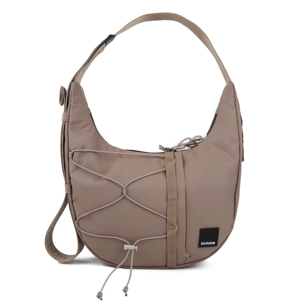 Taupe Kintobe crossbody bag with a curved silhouette, adjustable shoulder strap, and decorative drawstring accents.