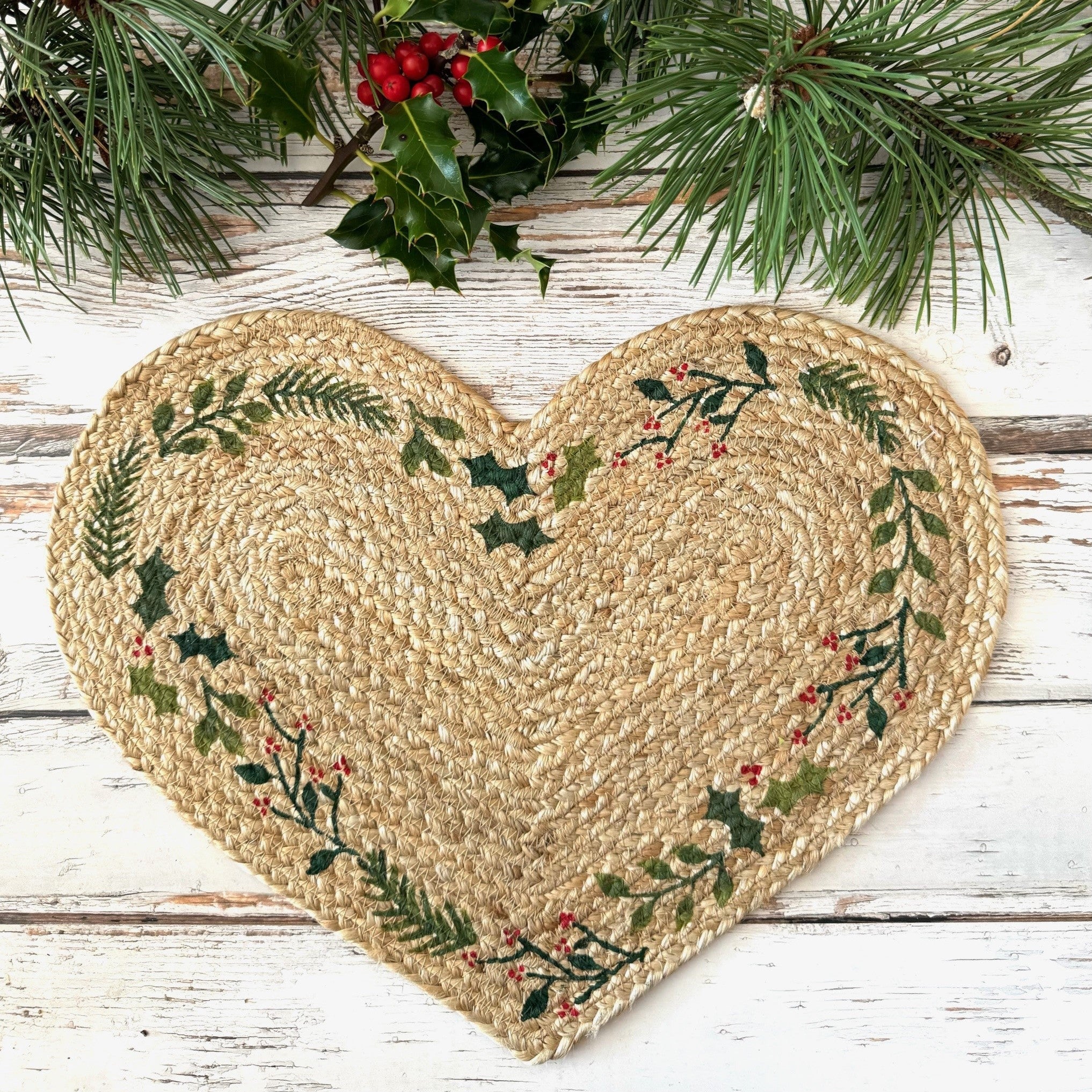 Heart-shaped jute mat with holly design on a wooden surface with pine branches.