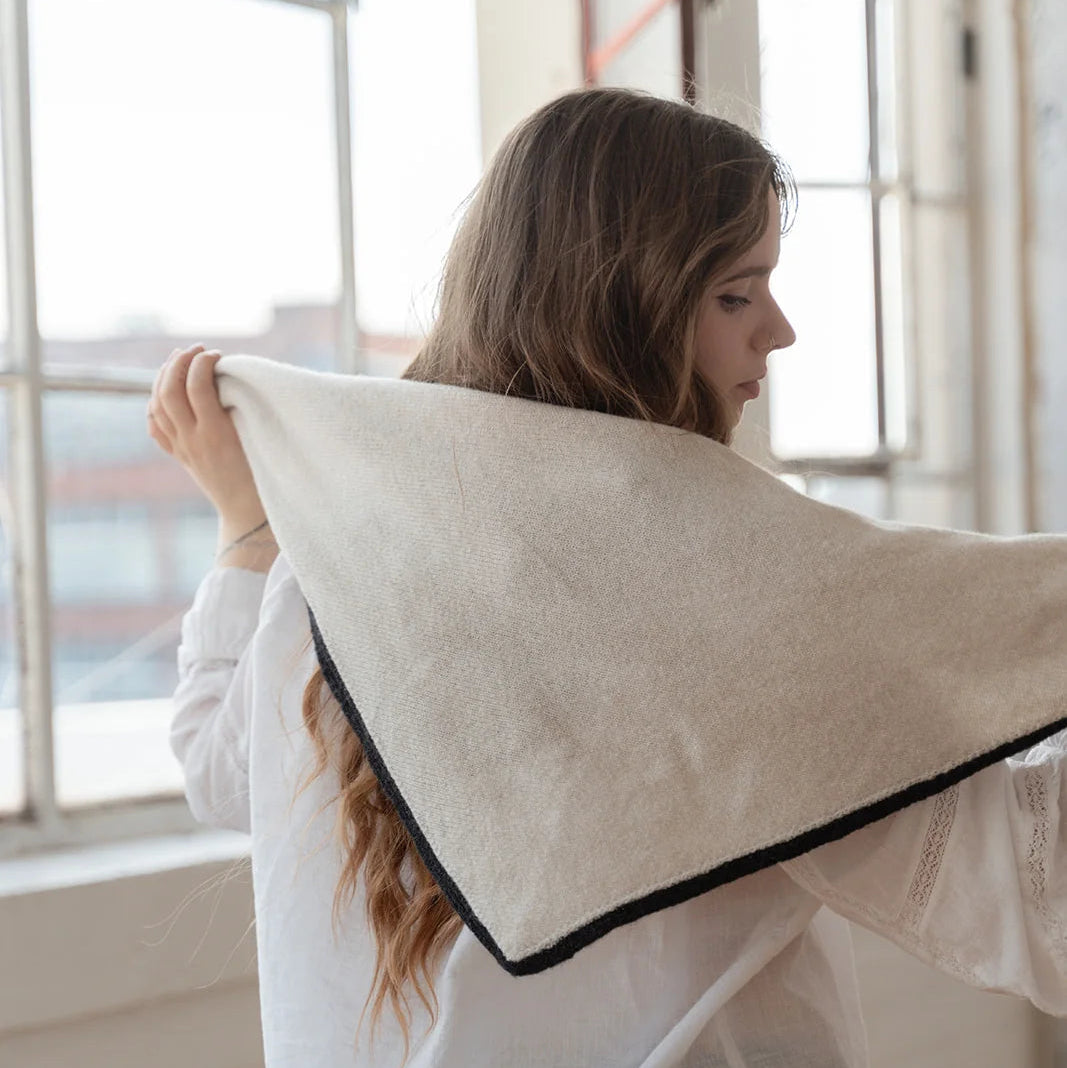 A woman in a white blouse and dark pants holds up a large square cream-colored textile, likely a scarf, shawl, or throw, which features a black piped border. She is photographed from behind in a bright, modern room with large windows.