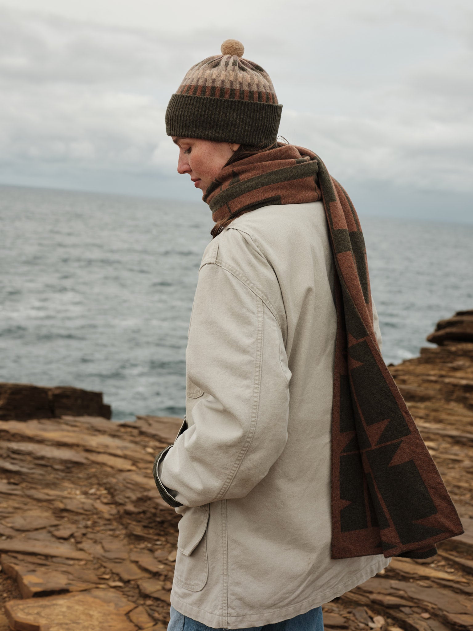 Person wearing a beige coat, brown beanie, and plaid scarf standing on a rocky coastline.