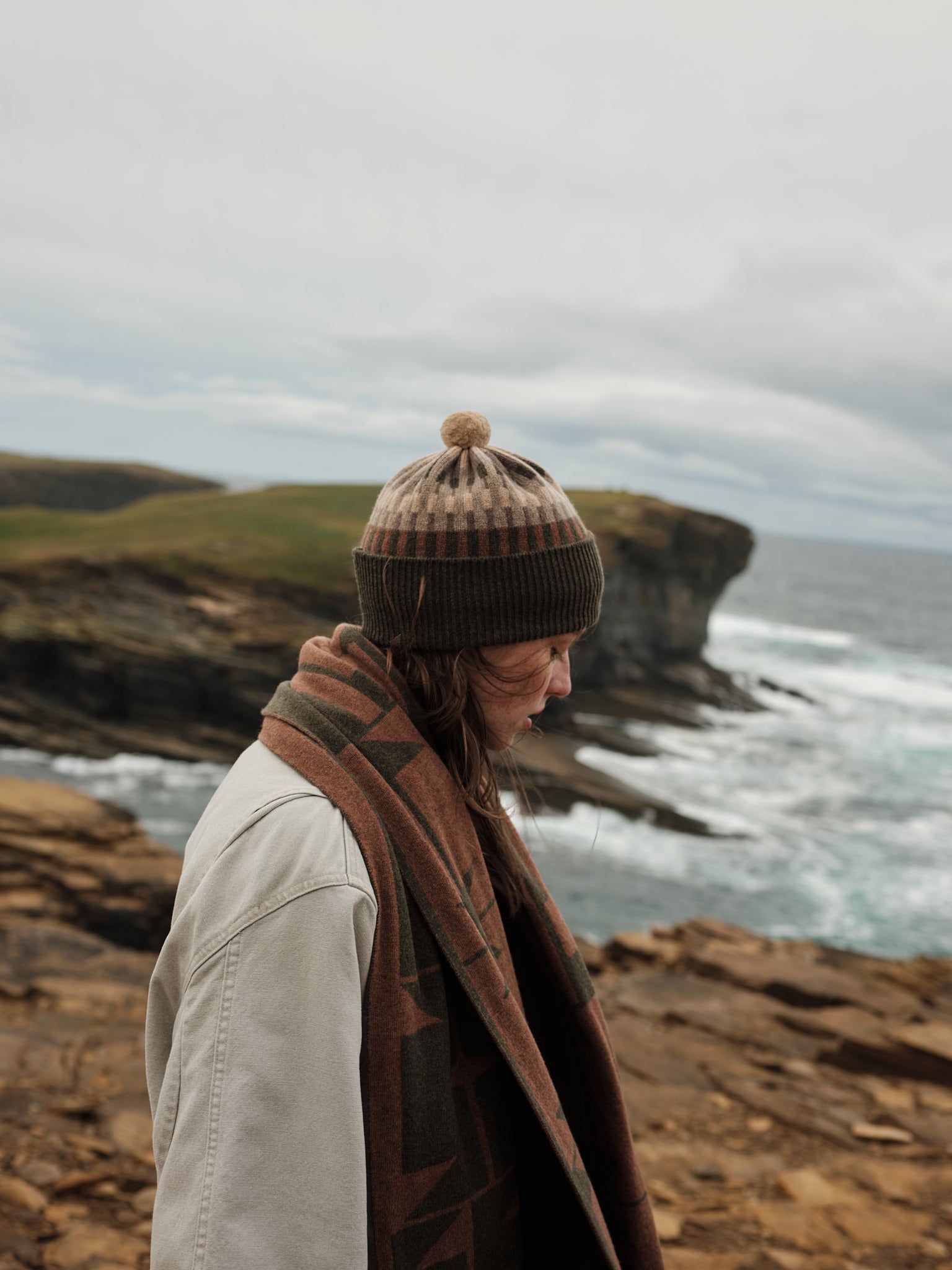 Person wearing a beanie and scarf standing on a cliff overlooking the ocean.