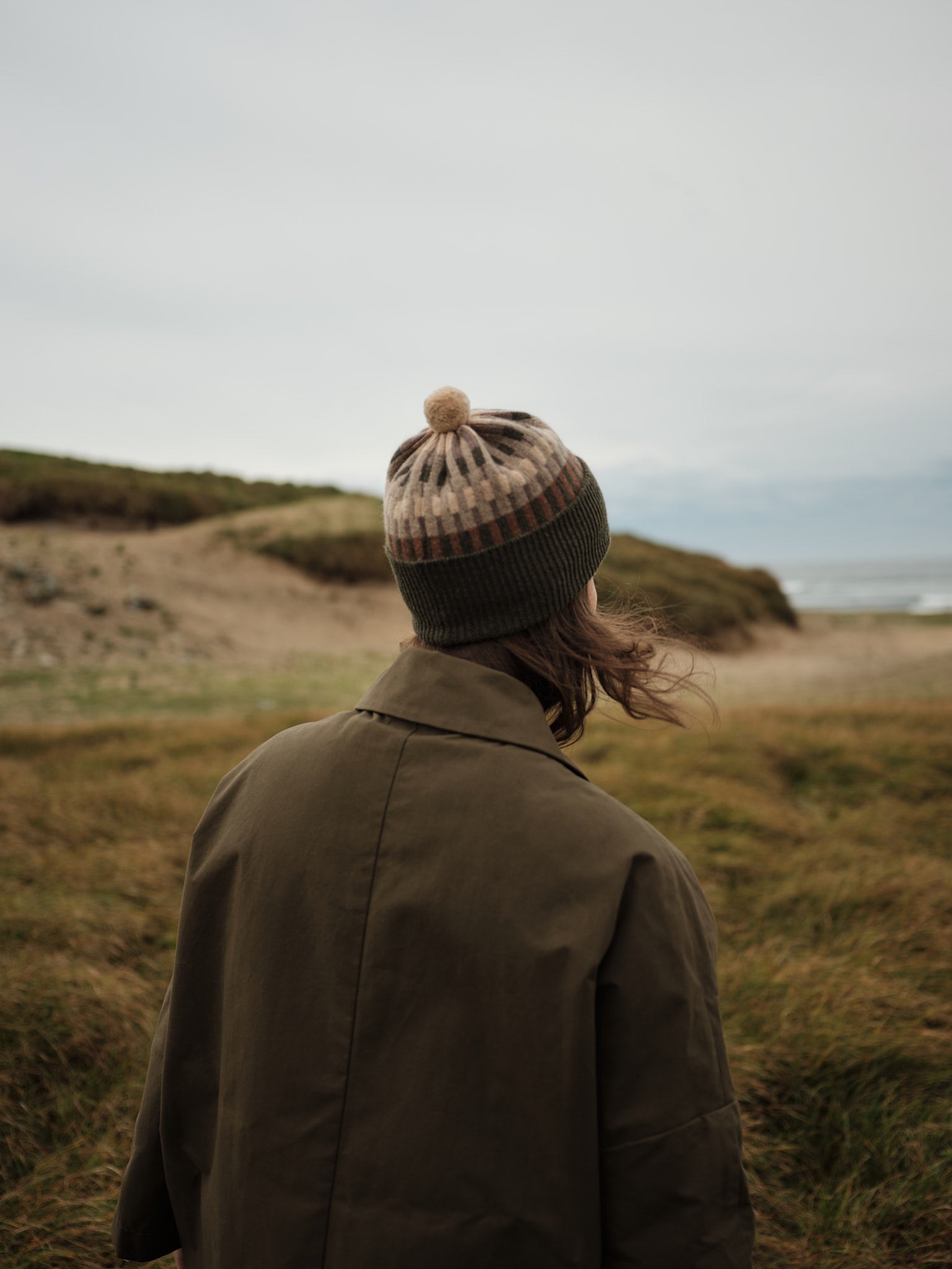 Person wearing a coat and knit hat standing in a grassy field with a scenic background
