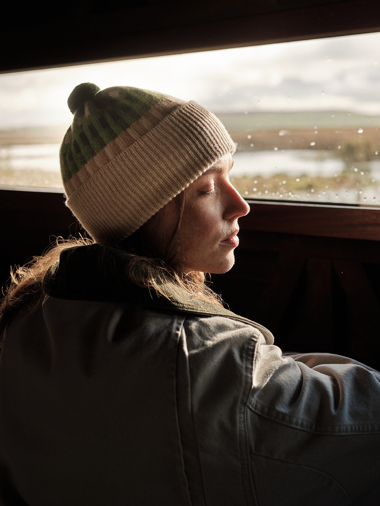 Person wearing a striped beanie and dark jacket looking out of a car window on a rainy day.