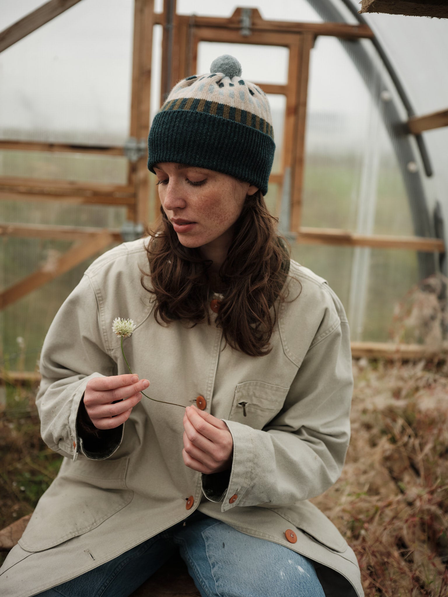 Person in a beige jacket and knit hat examining a plant inside a greenhouse.