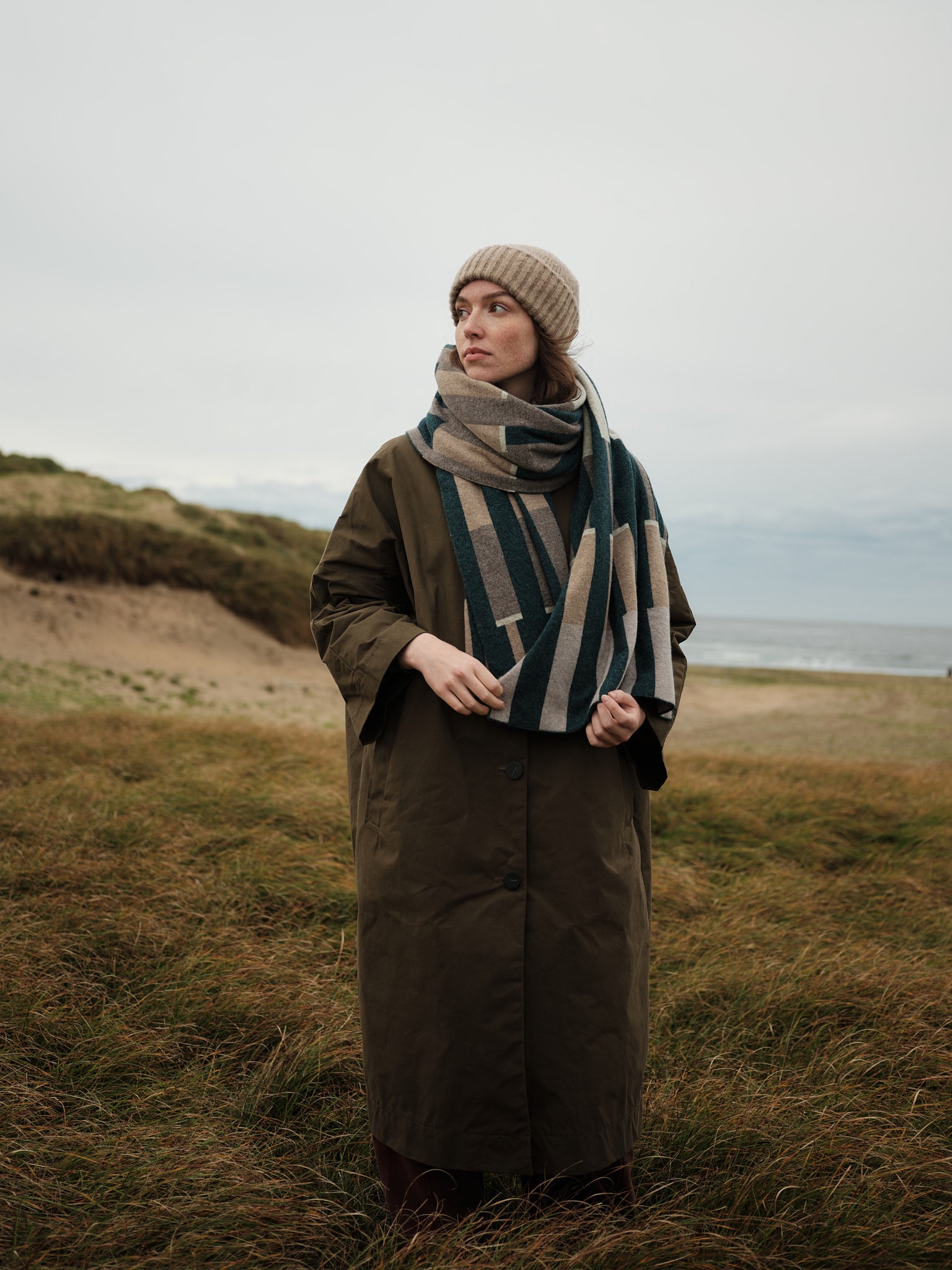 Person wearing a long coat and patterned scarf standing in a grassy field with a coastal background.