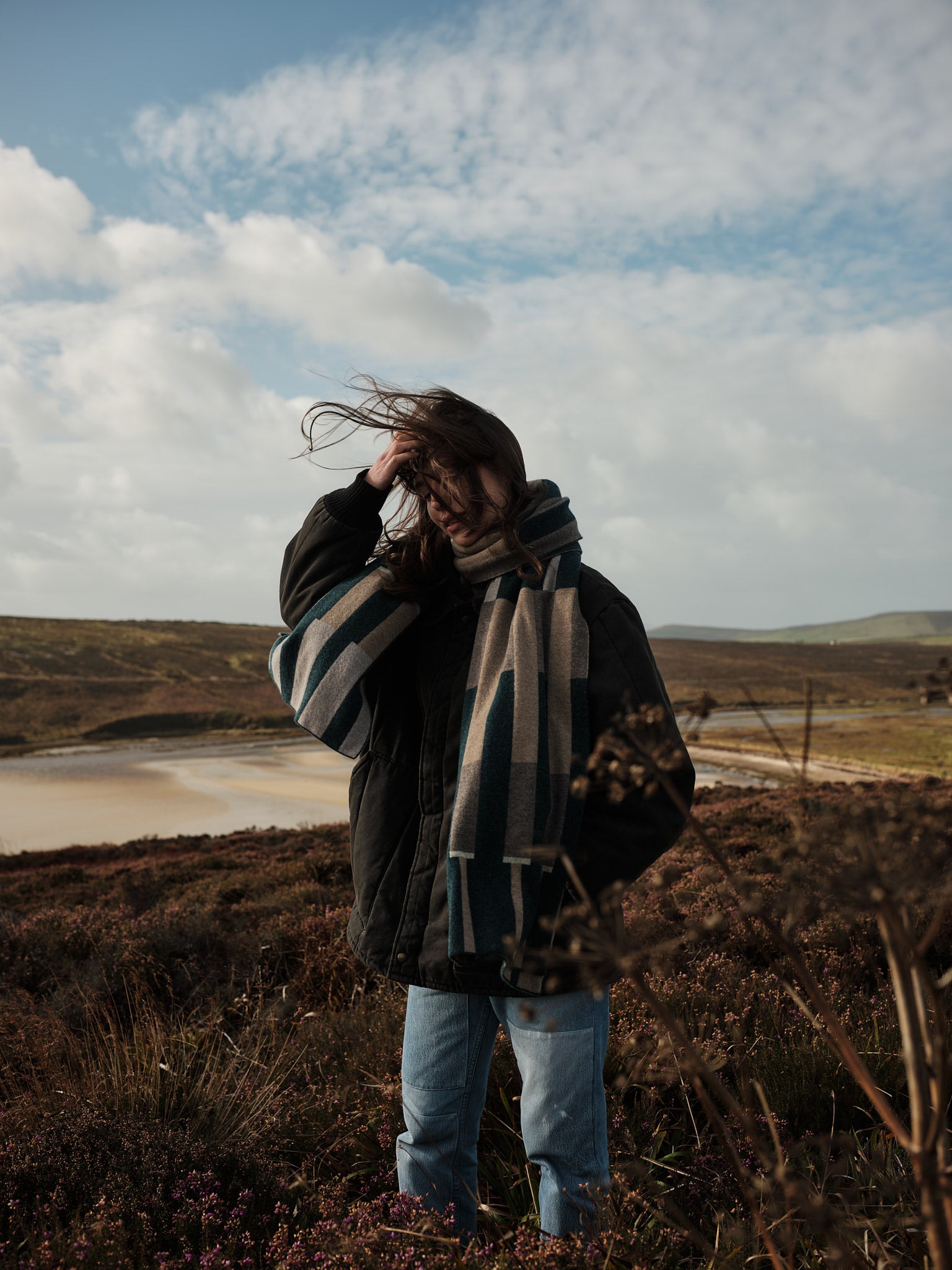 Person standing in a field with a scarf and coat, wind blowing hair