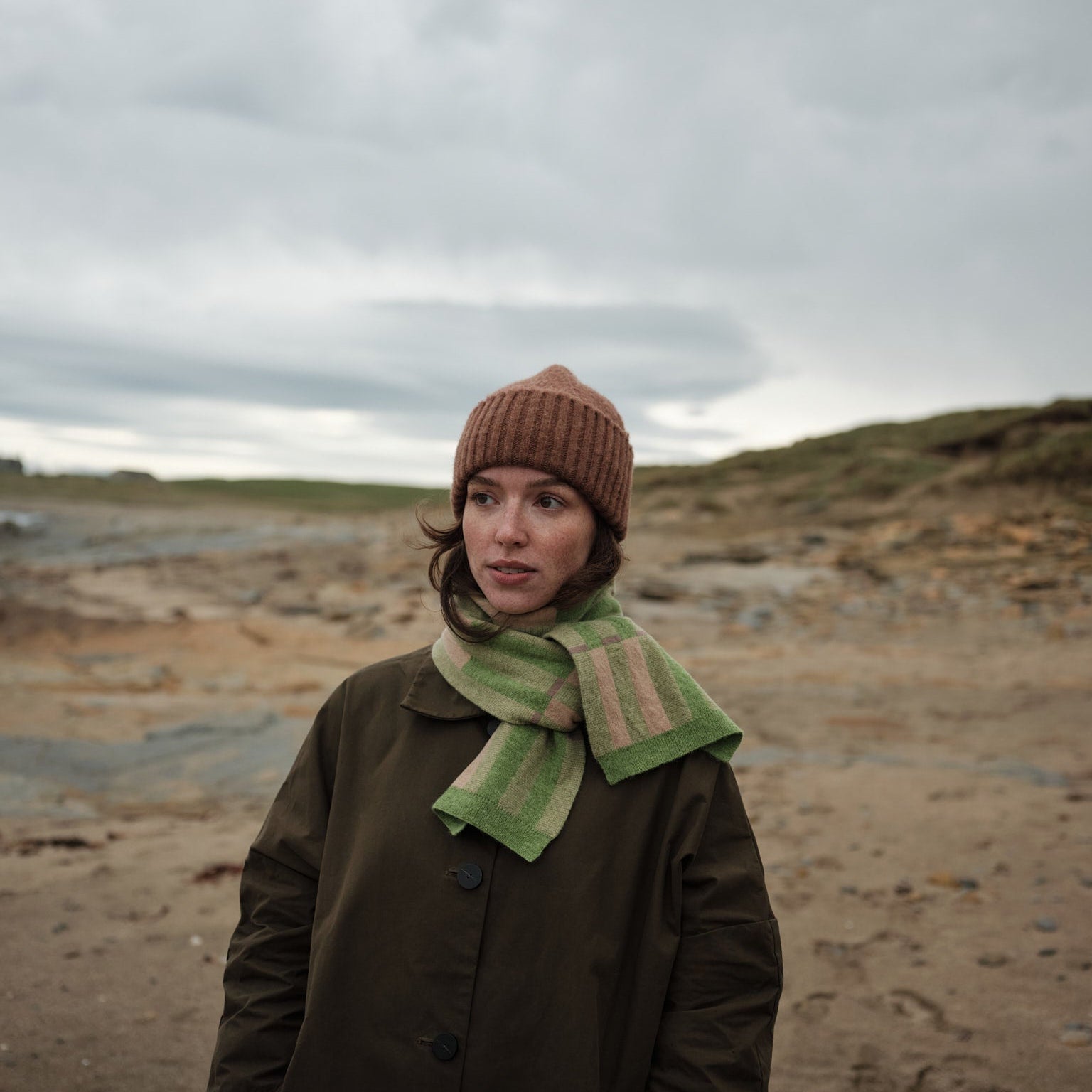 Person wearing a brown coat, green scarf, and knit hat standing on a beach with a cloudy sky.
