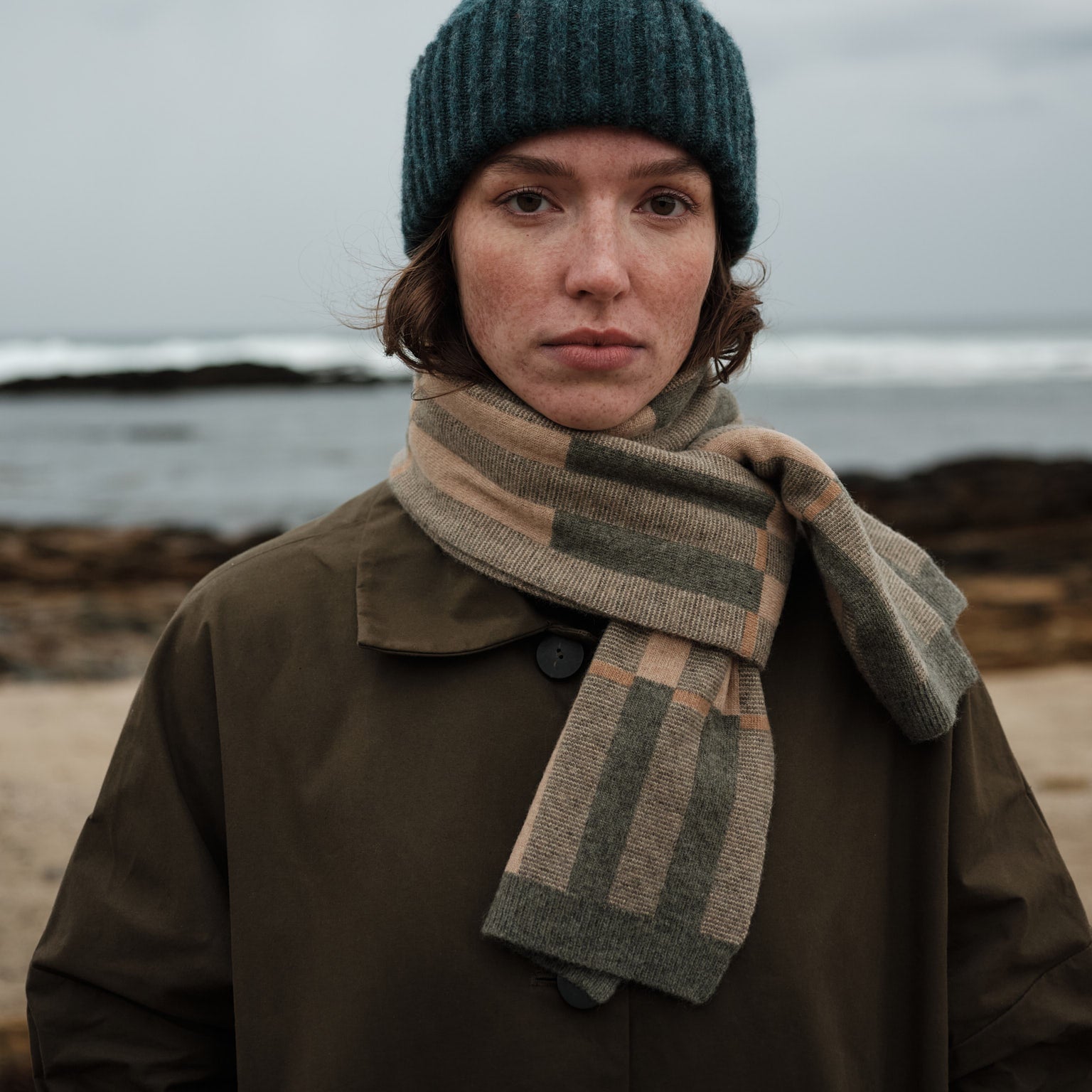 Person wearing a green knit hat and striped scarf standing on a beach with ocean in the background