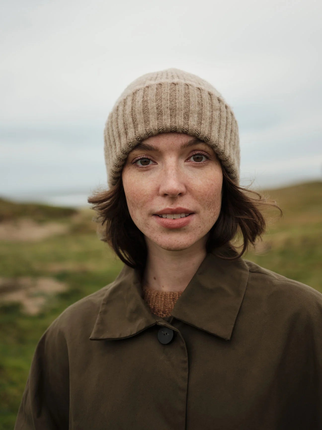 Person wearing a beige knit hat and brown coat in a natural setting
