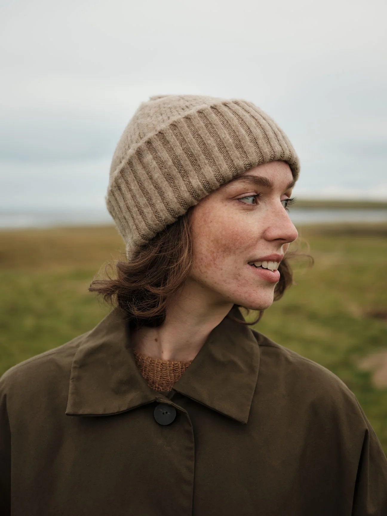 Person wearing a beige knit hat and brown coat in a field