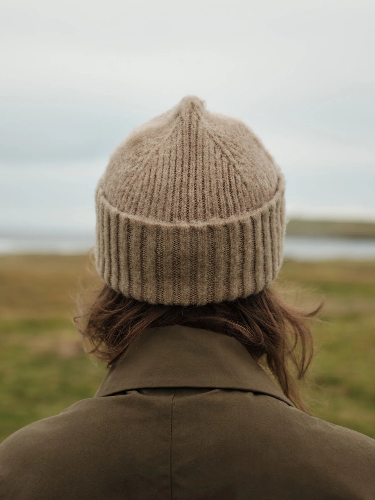 Person wearing a beige knit beanie and coat standing in a grassy field with a blurred background