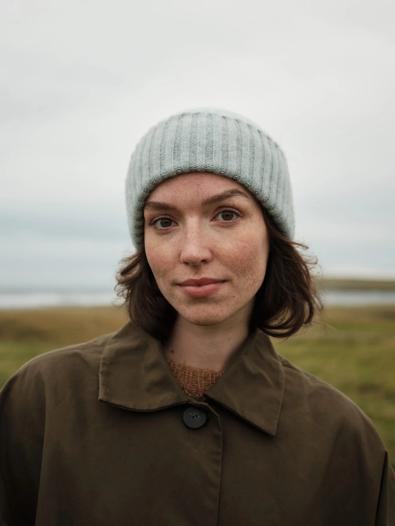 Person wearing a light gray knit hat and brown coat in a natural setting