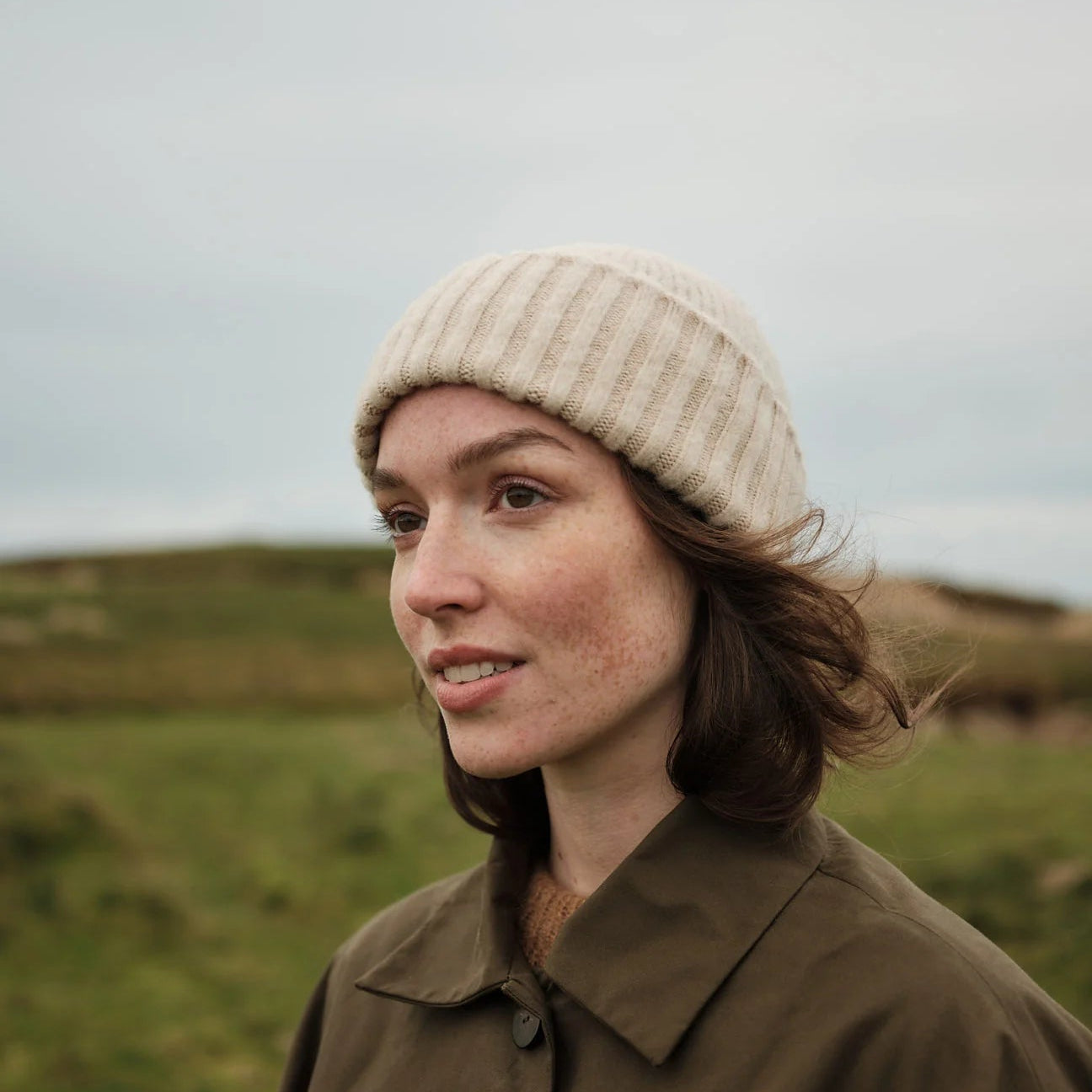Woman wearing a beige knit hat and brown coat standing in a grassy field.