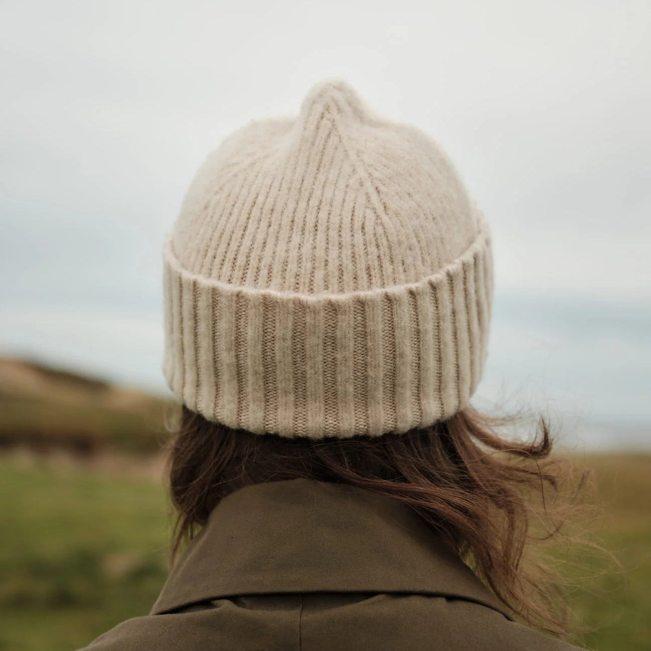 Person wearing a beige knit beanie and brown coat standing in a field with a blurred background