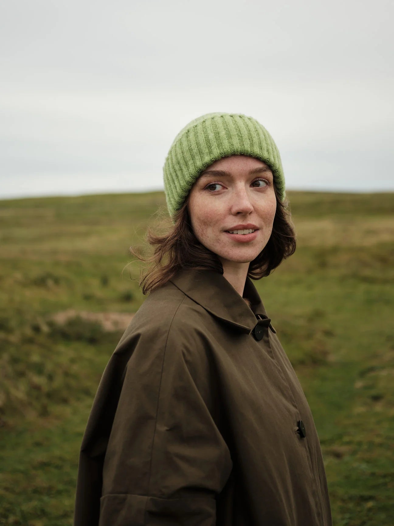 Person wearing a green knit hat and brown coat standing in a grassy field.