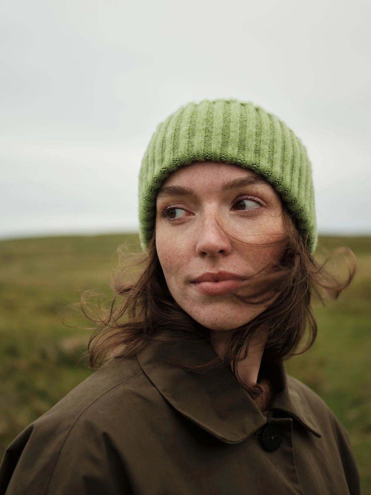 Person wearing a green knitted hat and brown coat in a grassy field