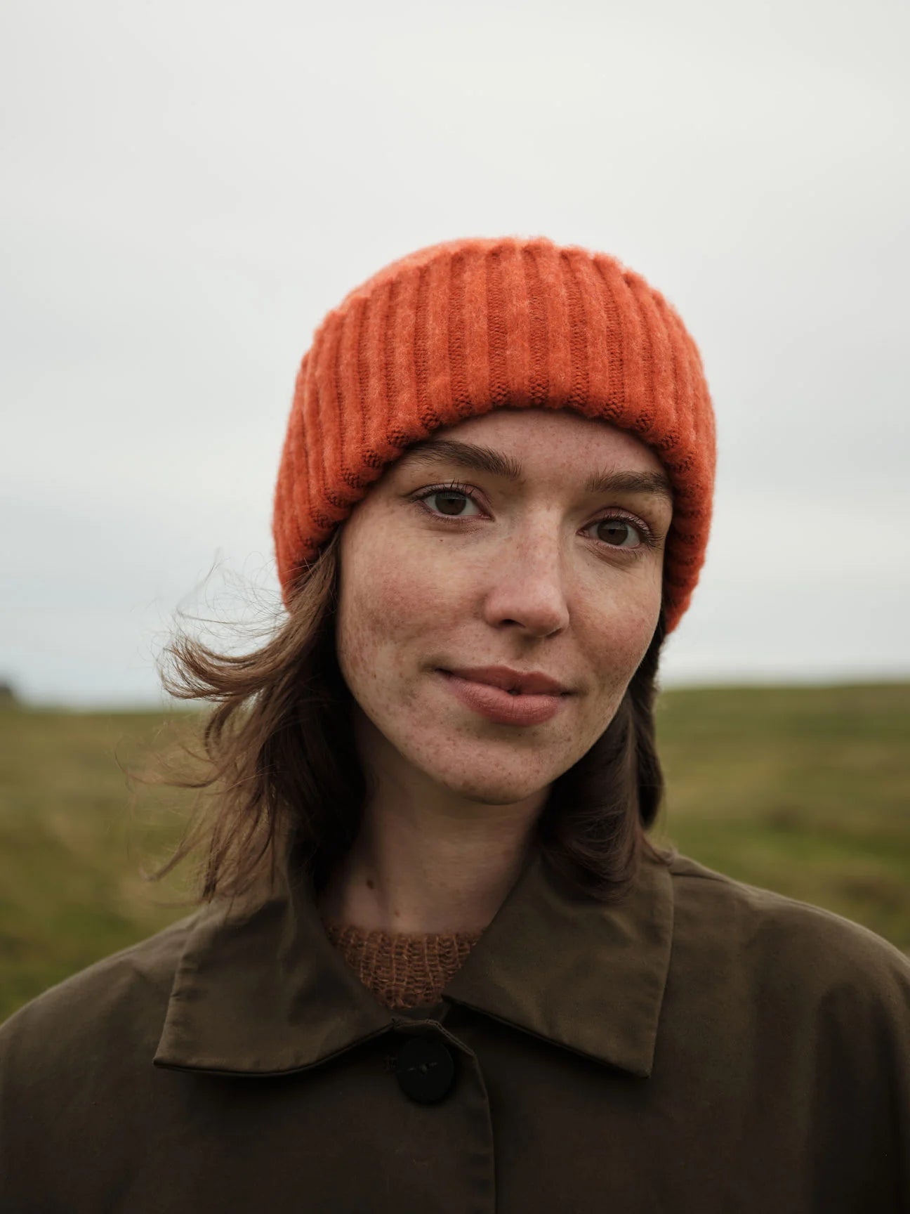 Person wearing an orange knit hat and brown coat in a natural setting