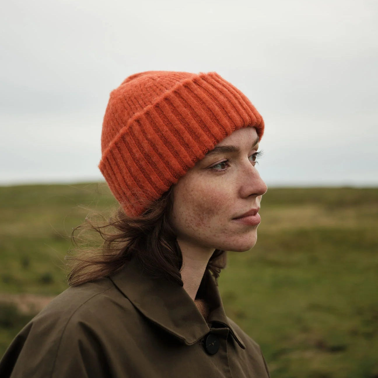 Person wearing an orange knit hat and brown coat standing in a grassy field.