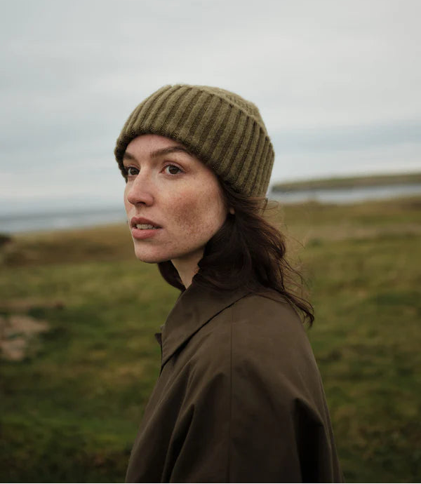 Woman wearing a green knitted beanie and brown coat standing in a grassy field with a body of water in the background.