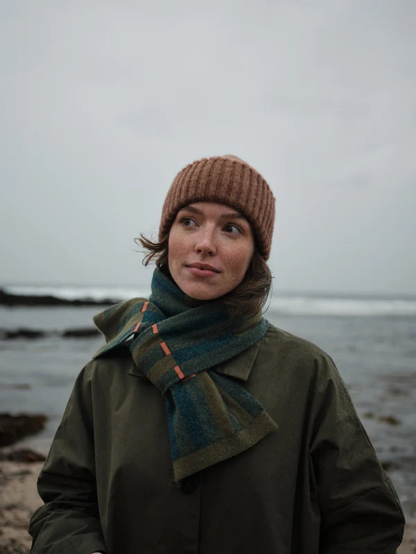 Person wearing a brown knit hat and green coat with a scarf, standing on a beach.