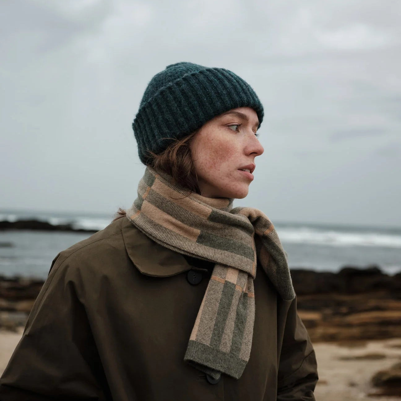 Person wearing a beanie and scarf standing on a beach with a cloudy sky.