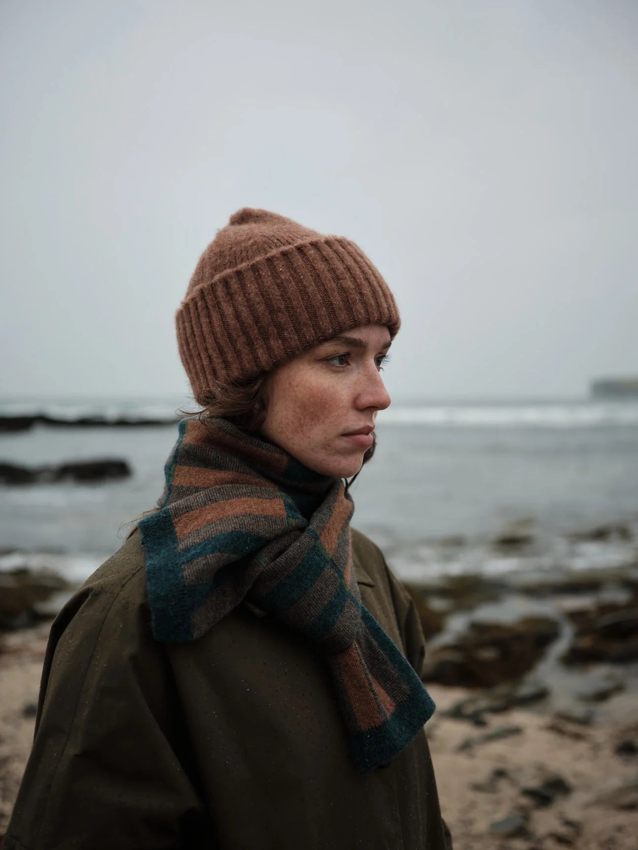 Person wearing a brown knit hat and scarf standing on a beach with a gray sky.