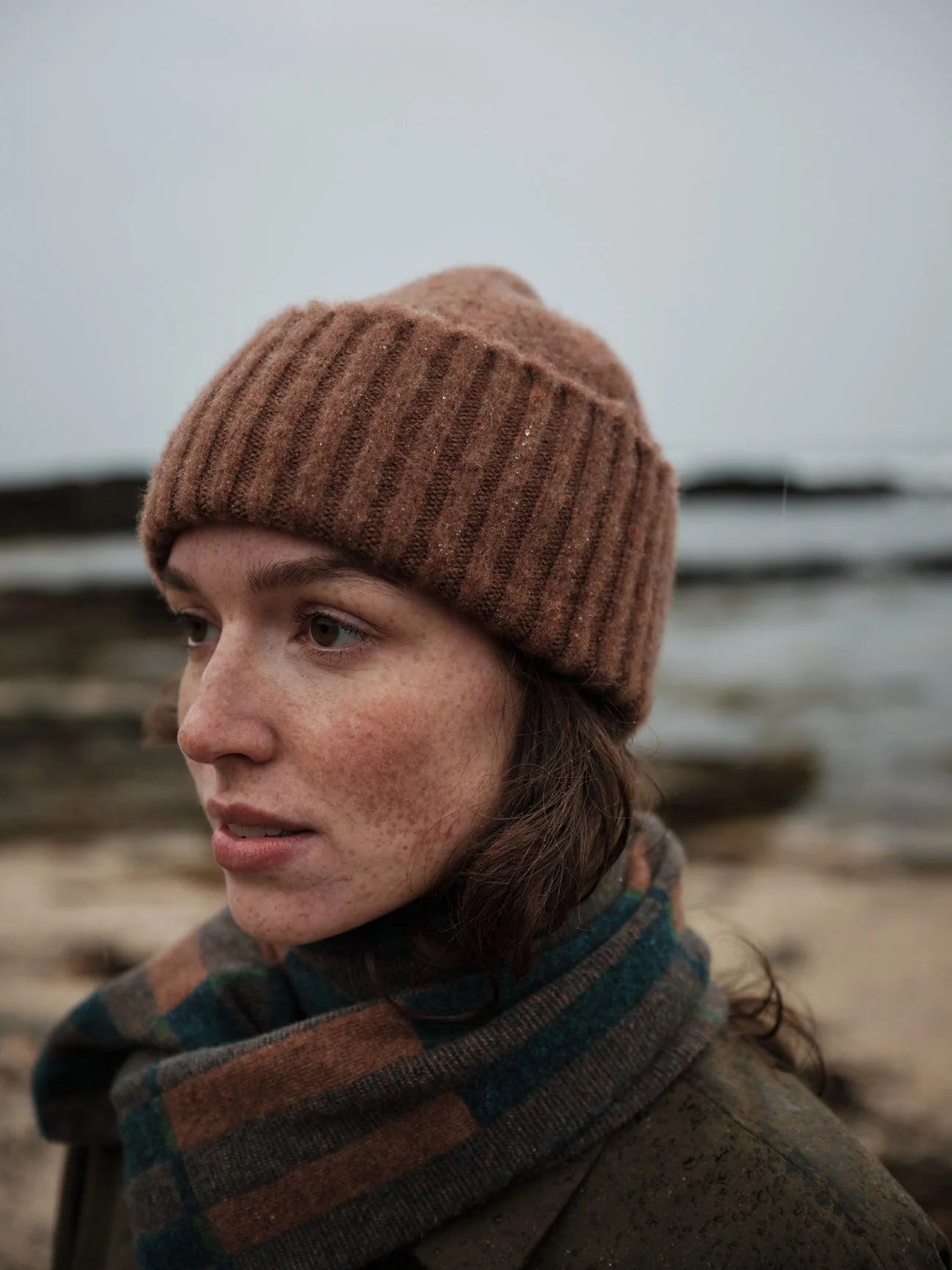 Person wearing a brown knit beanie and plaid scarf by a body of water.