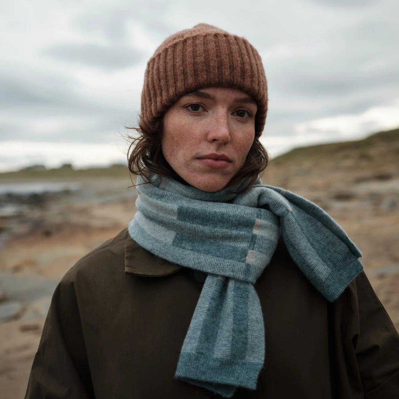 Person wearing a brown knit hat and blue scarf in a barren landscape