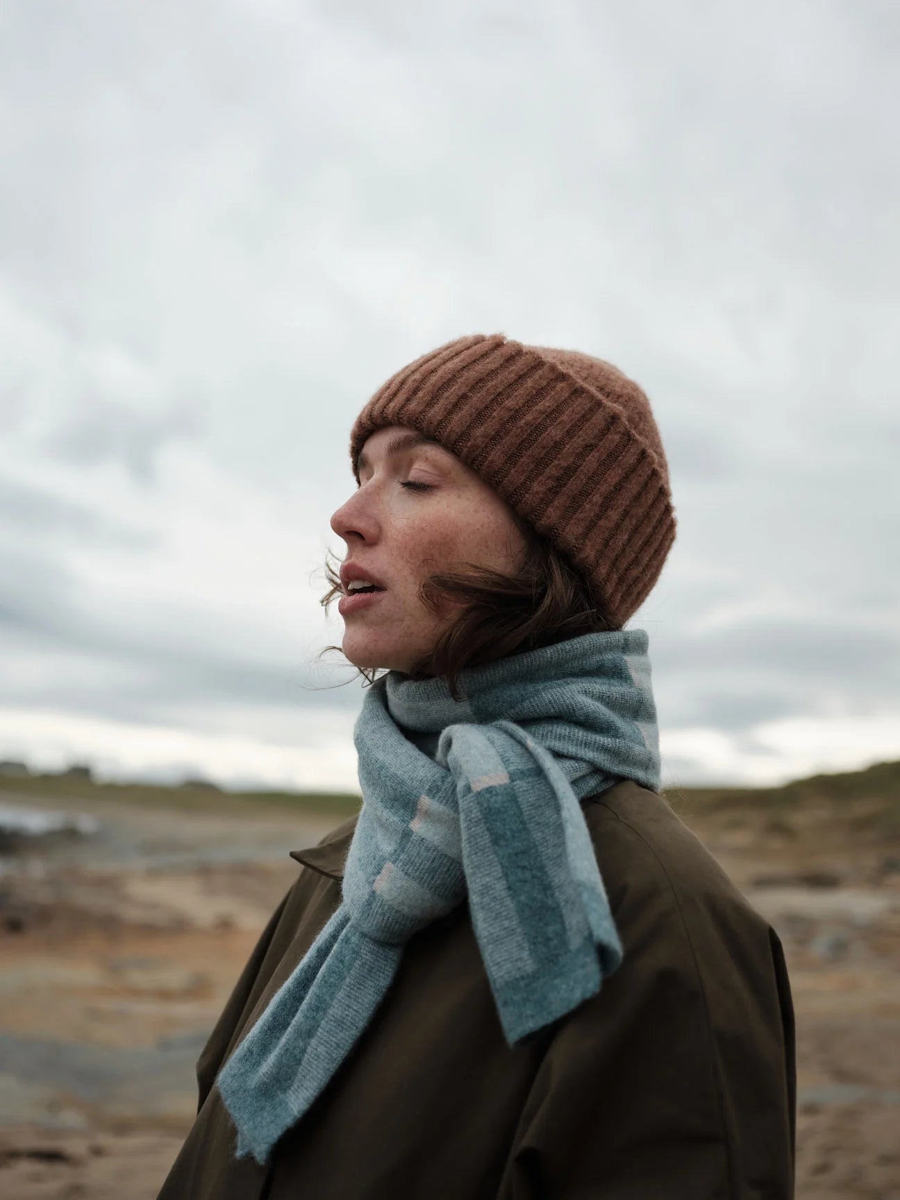 Person wearing a brown knit hat and blue scarf in a barren landscape