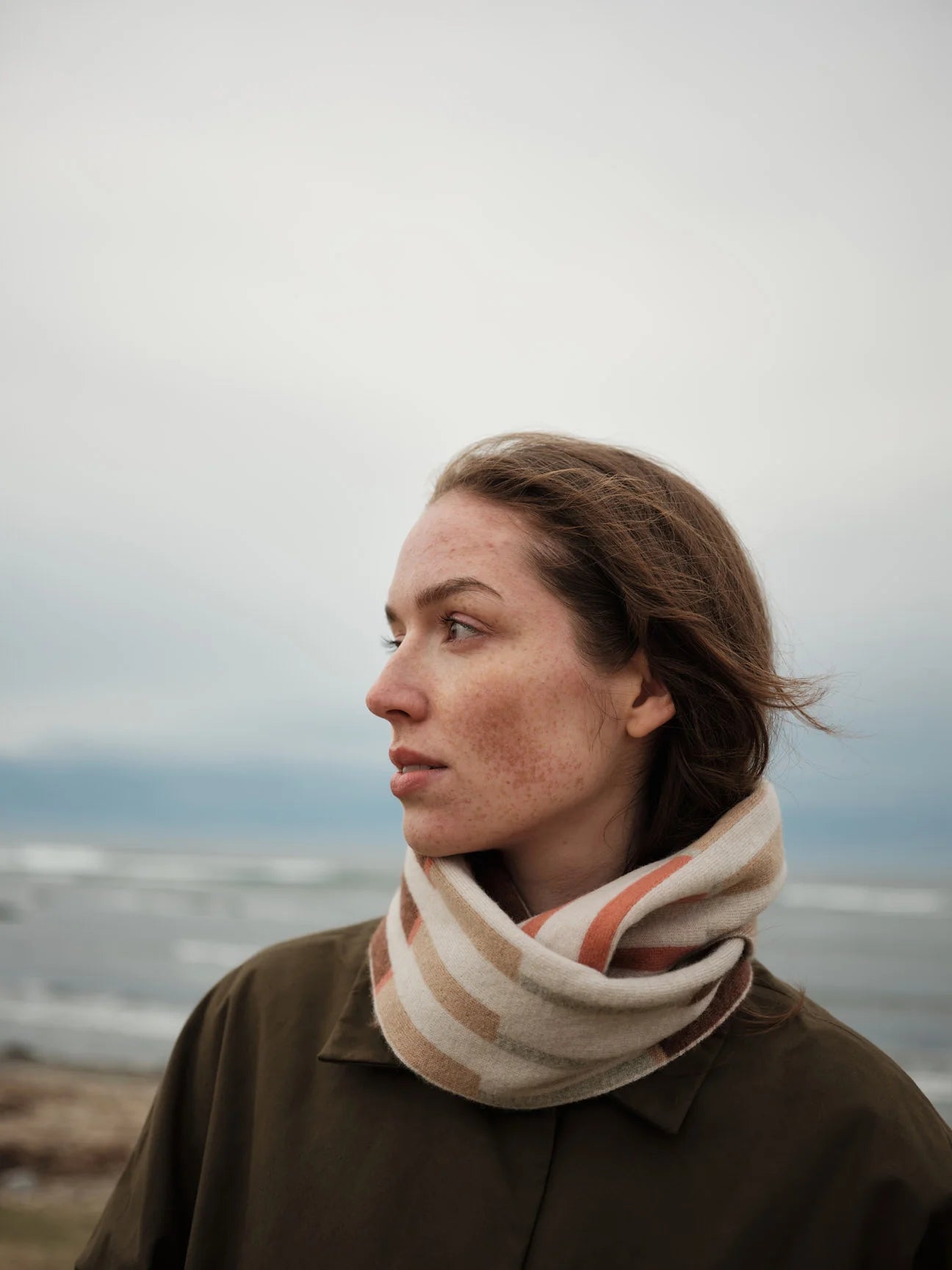 A woman in profile wearing a thick beige and orange striped knit scarf and a dark olive brown jacket, set against a soft-focus coastal background.