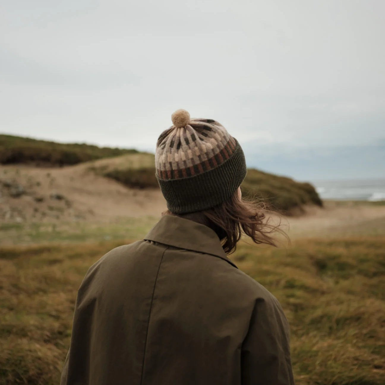 Person wearing a beanie and coat standing in a grassy field with a scenic background