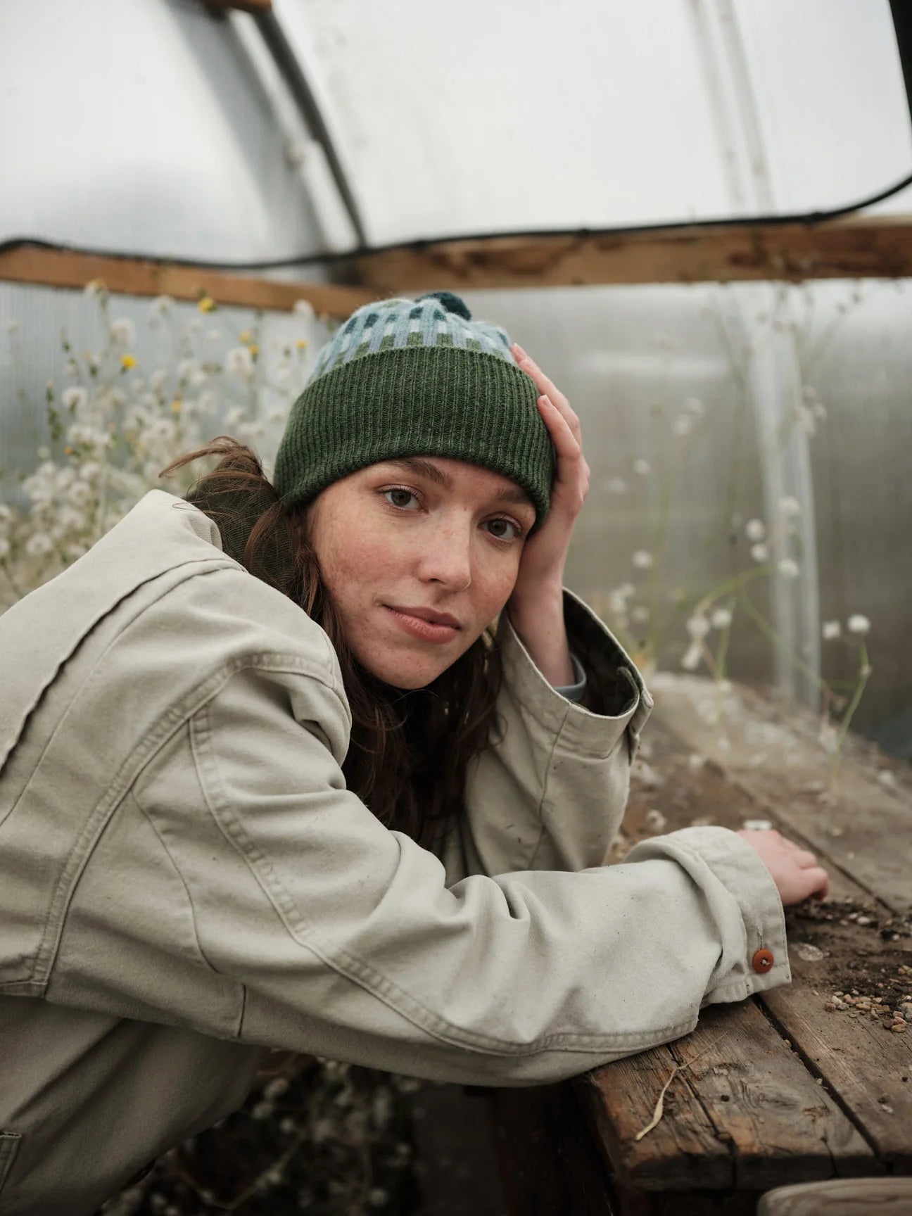 Person wearing a green beanie and beige jacket in a greenhouse setting