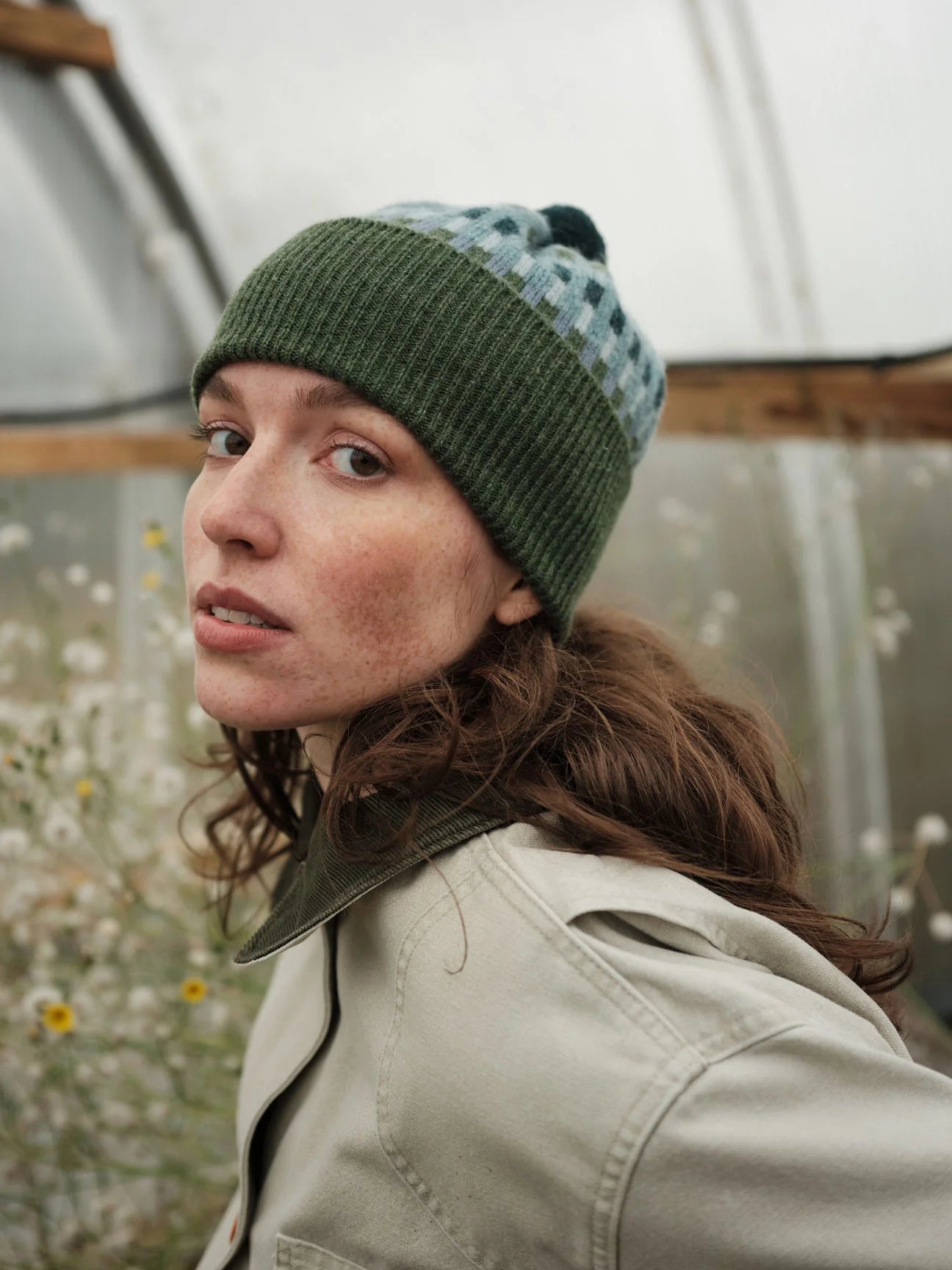 Person wearing a green knit beanie with a checkered pattern inside, standing in a greenhouse.