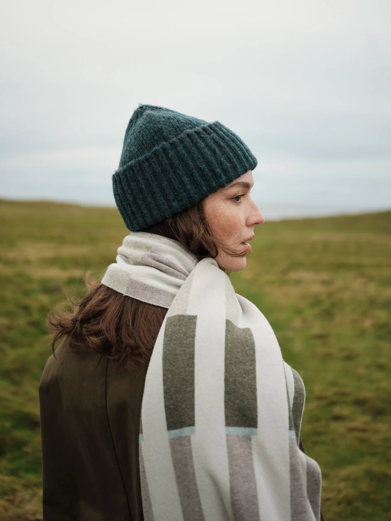 Person wearing a green knit beanie and a patterned scarf in a grassy field
