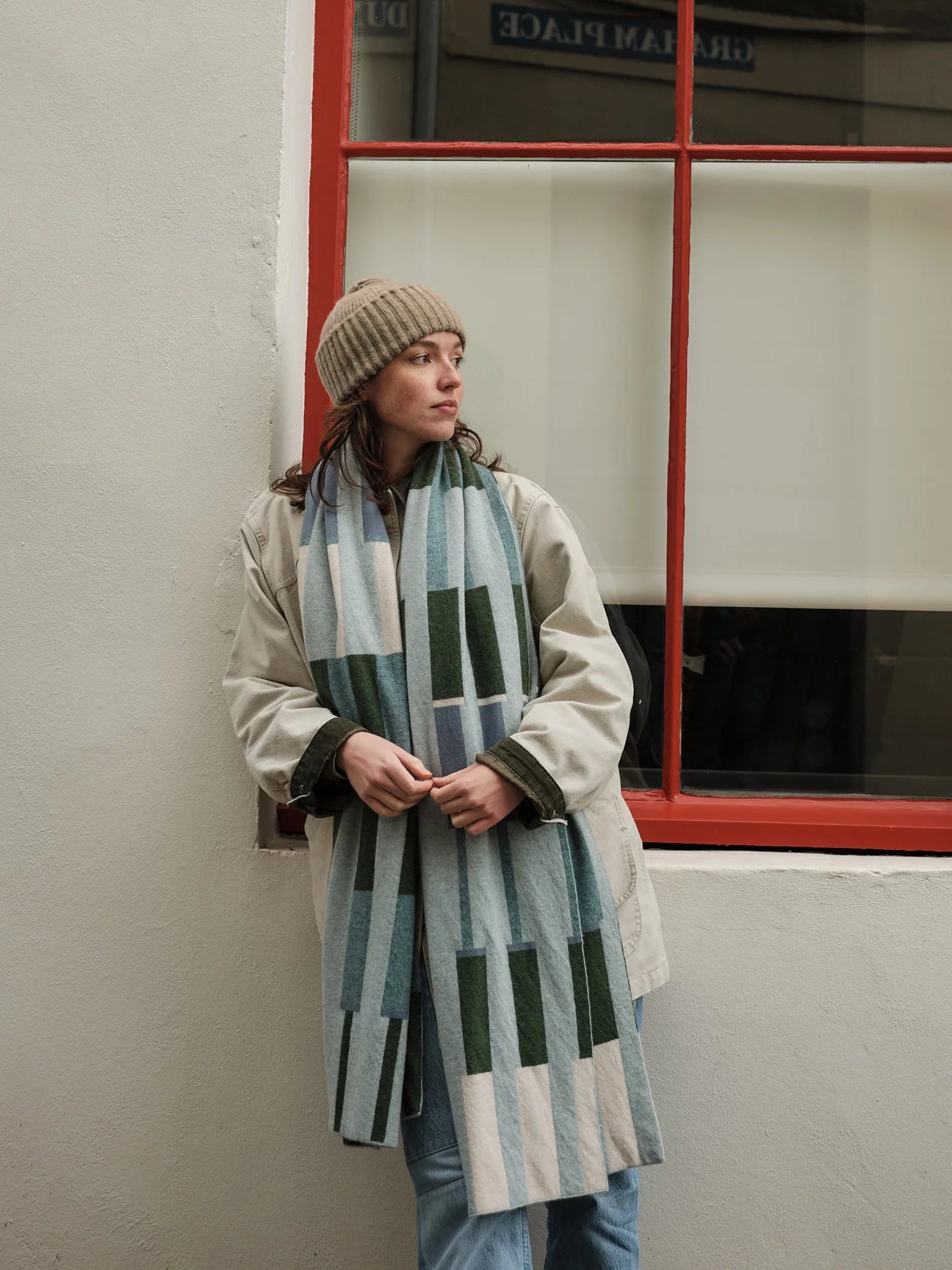 Person wearing a long, patterned scarf standing against a wall with a red-framed window.
