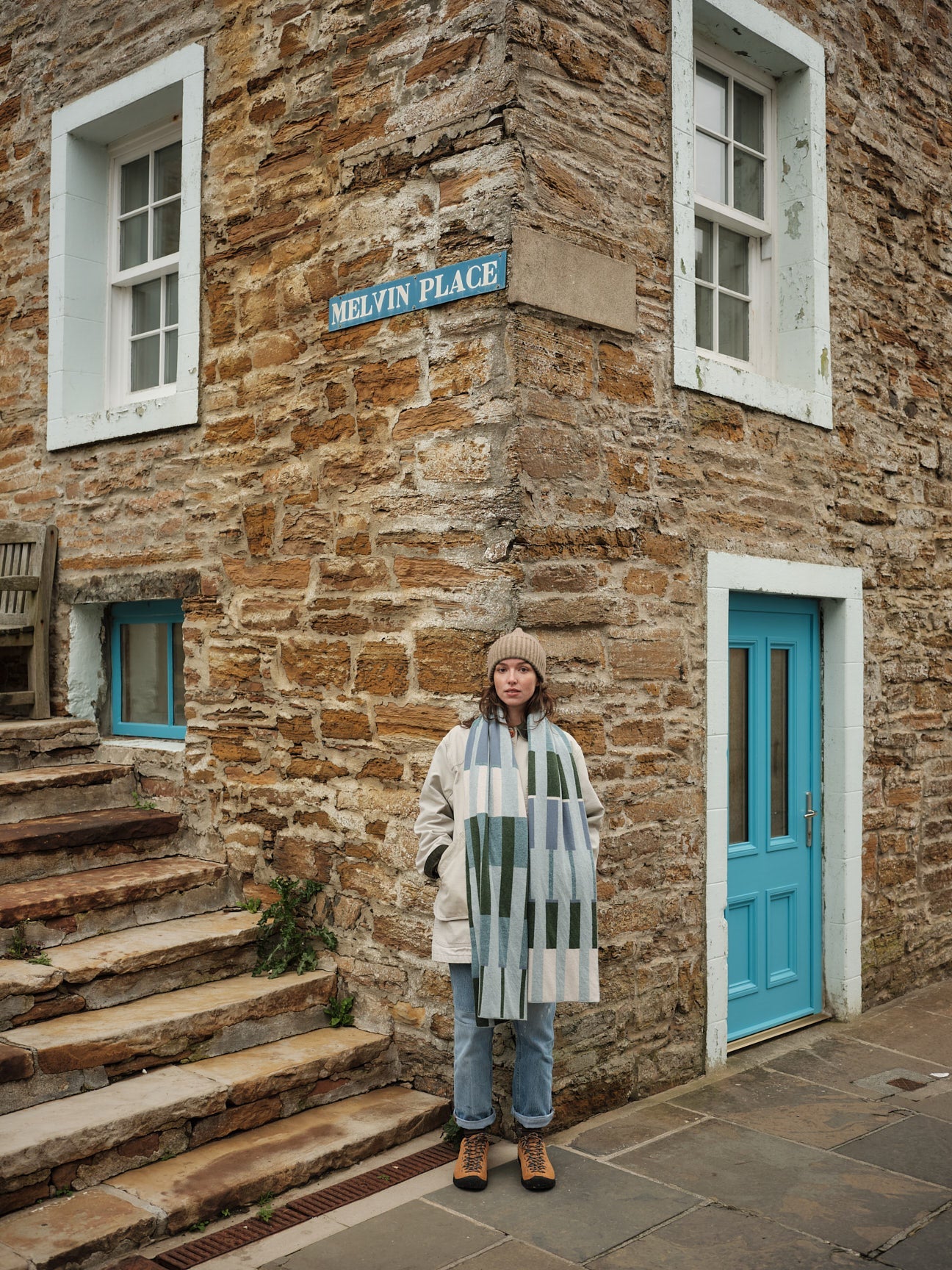 Person standing in front of a stone building with a blue door and sign reading 'Melvin Place'.