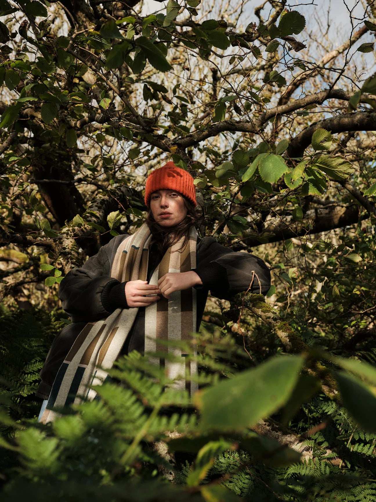 Person wearing a red beanie and checkered scarf standing among trees and foliage.