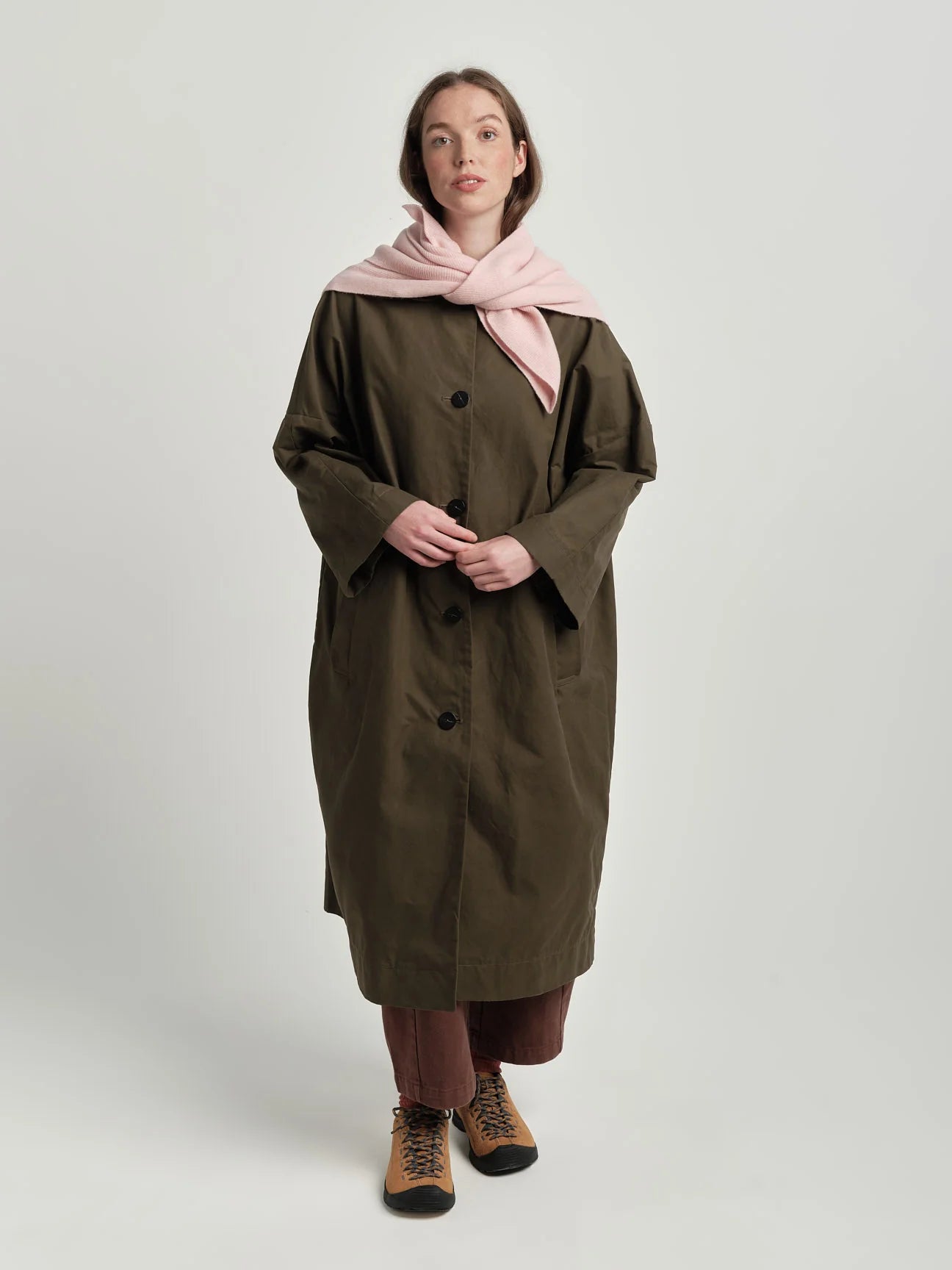 Woman wearing a long olive green coat with a pink scarf on a white background