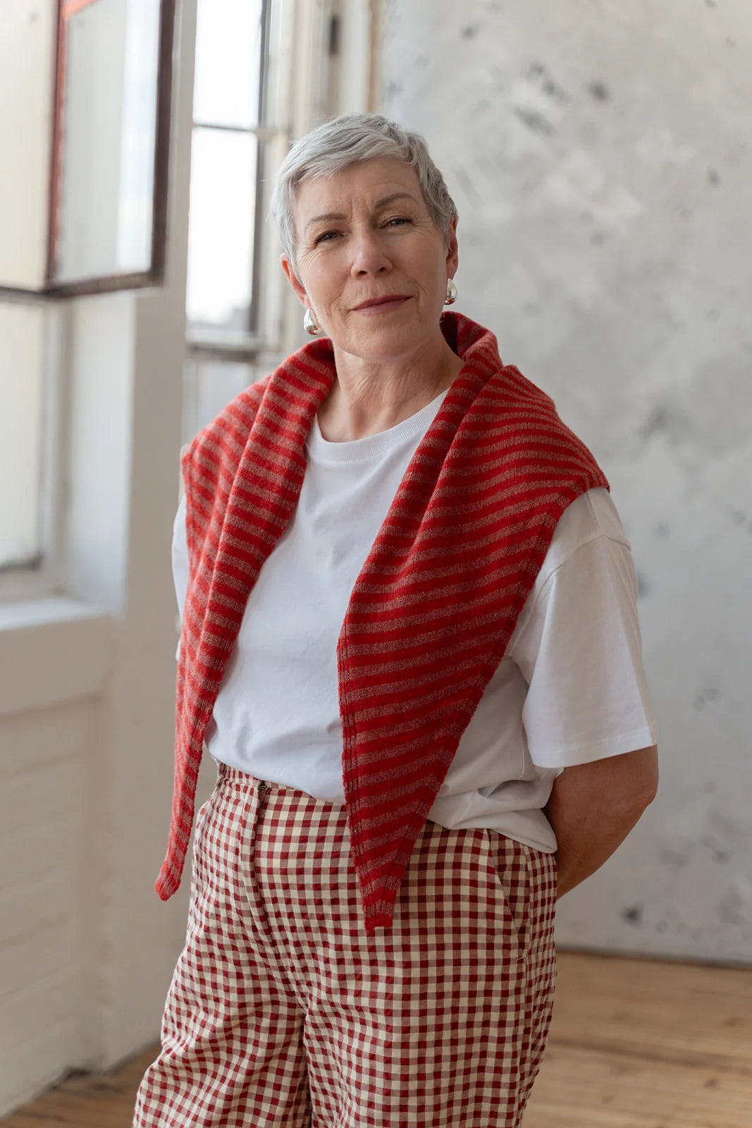 A woman with short gray hair stands indoors wearing a red-and-white striped knit shawl draped over her shoulders, a white T-shirt, and red-and-white gingham pants.
