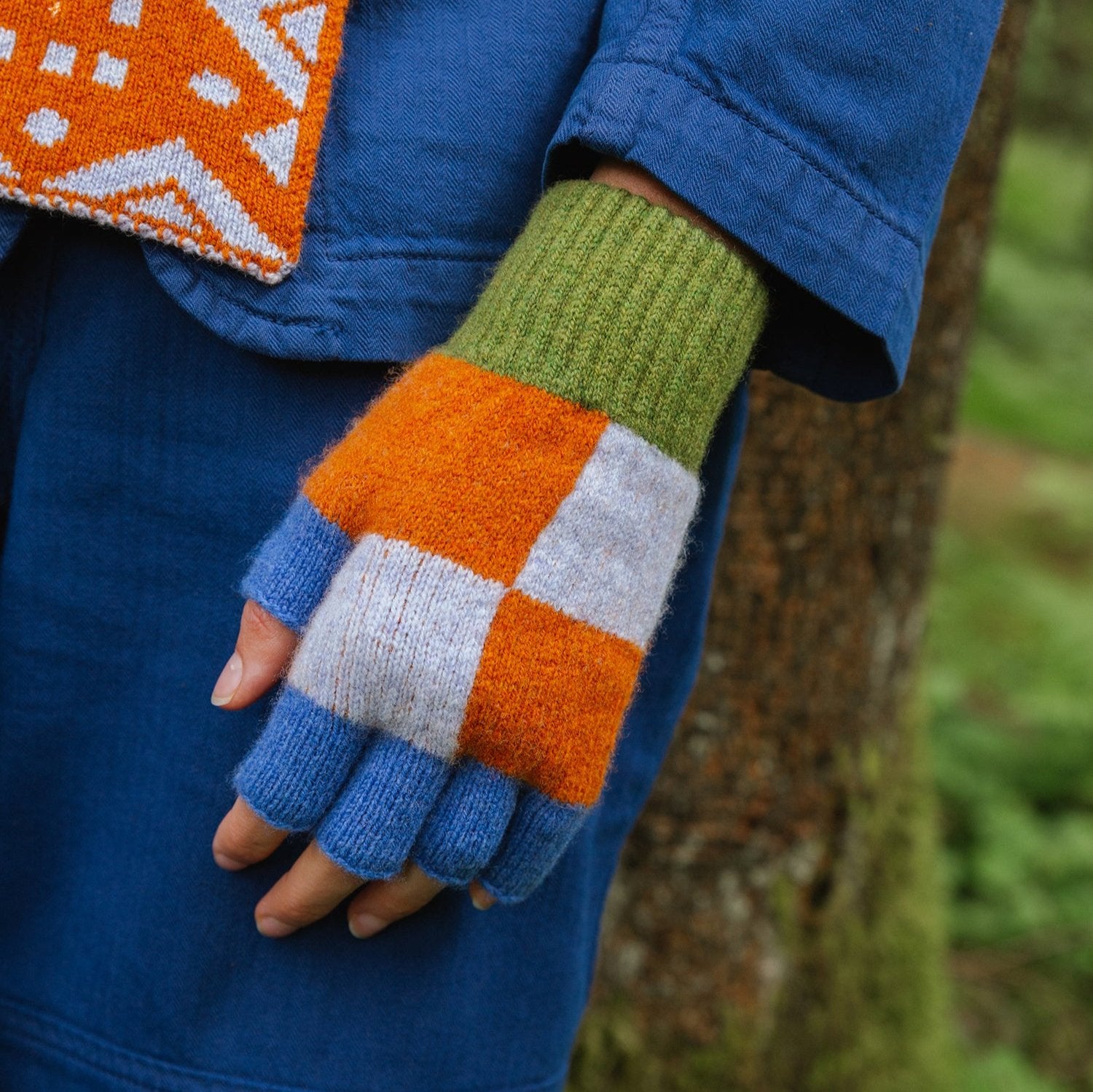 Hand wearing fingerless gloves with orange, blue, white, and green color-blocked knit pattern.