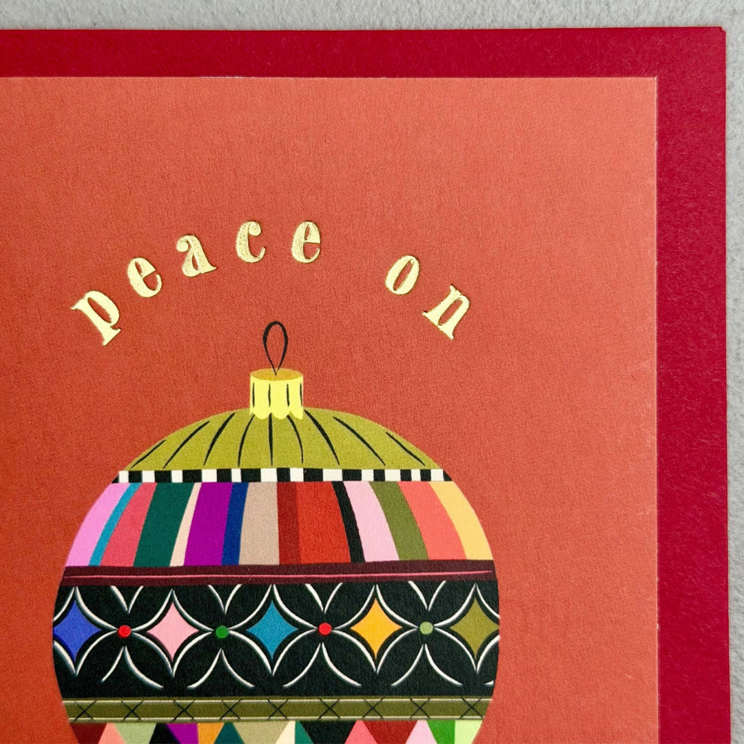 A close-up of a greeting card featuring the words 'peace on' in gold lettering above a colorful patterned Christmas ornament. The card has a warm coral-red background and a darker red border, photographed against a light gray surface.