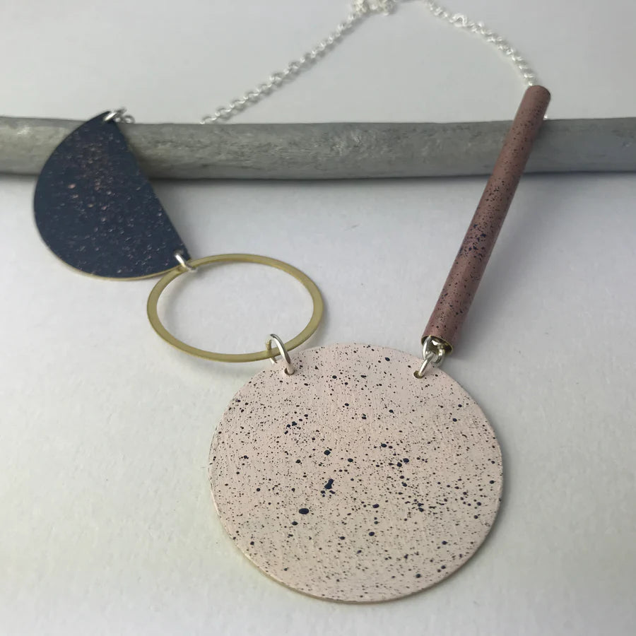 Geometric statement necklace with speckled pink and dark blue/gray semi-circular and circular elements, brass ring, and