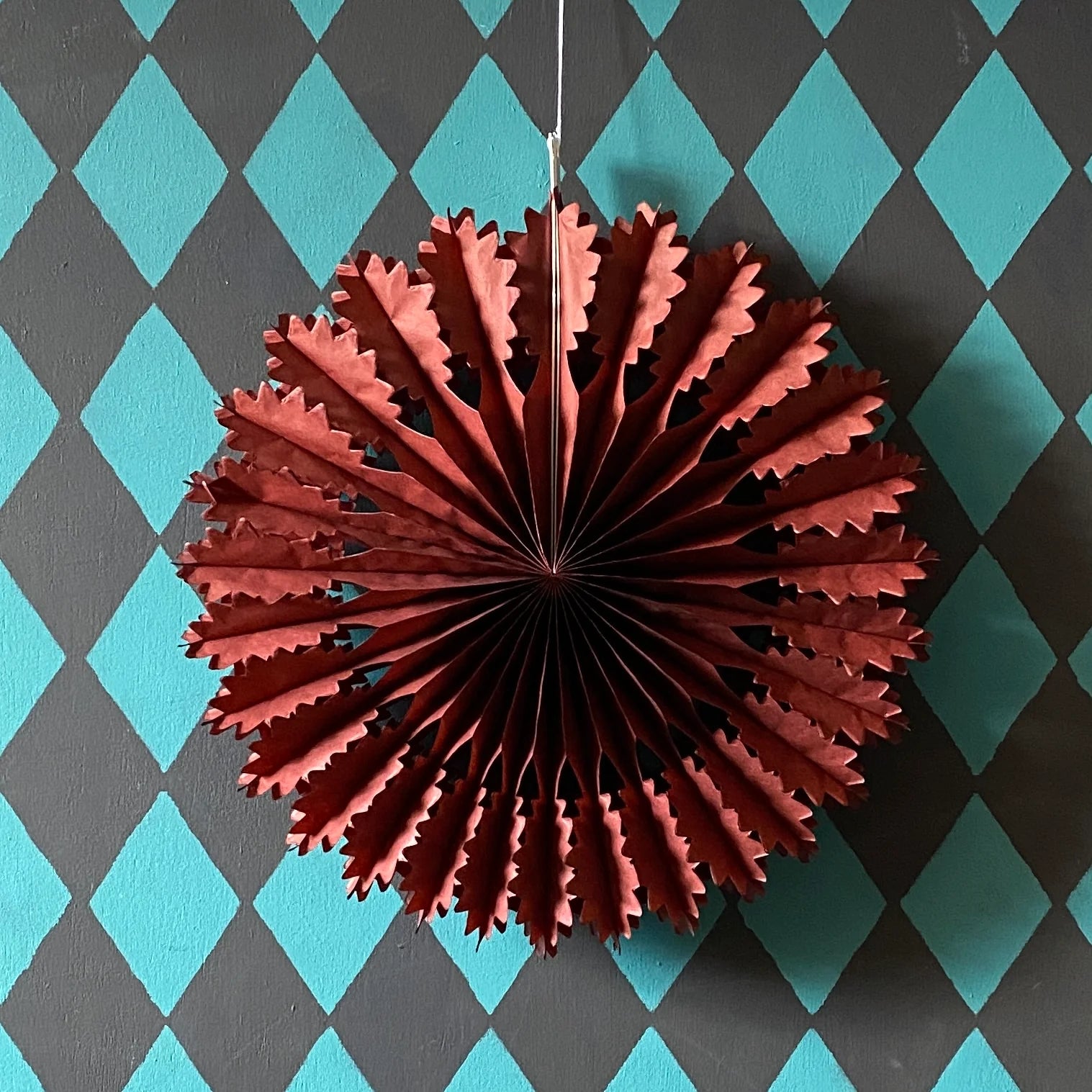 Russet-colored paper fan decoration hanging against a teal and gray diamond patterned wall.