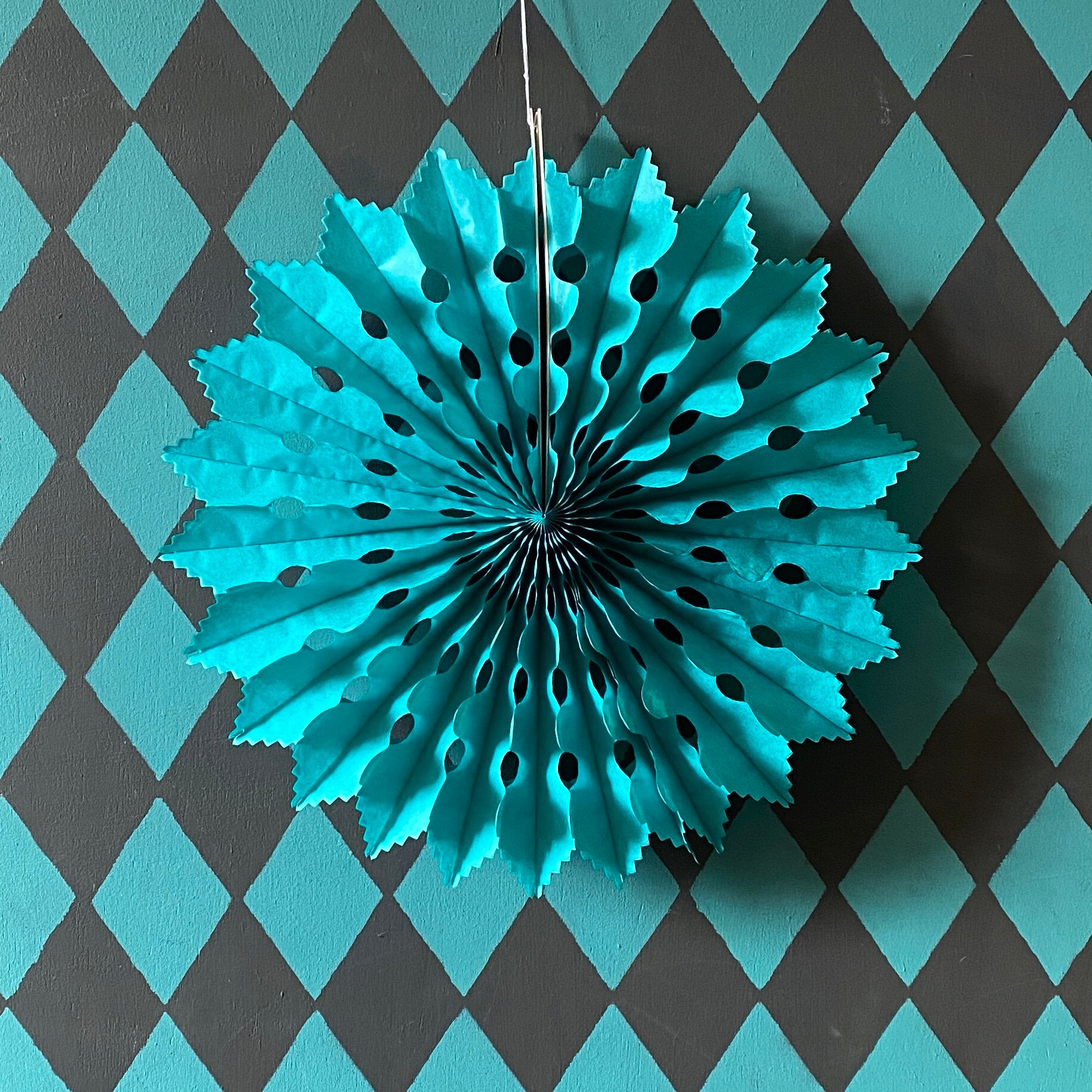 Teal paper fan decoration with scalloped edges and cut-out holes, hanging against a dark gray and teal harlequin diamond