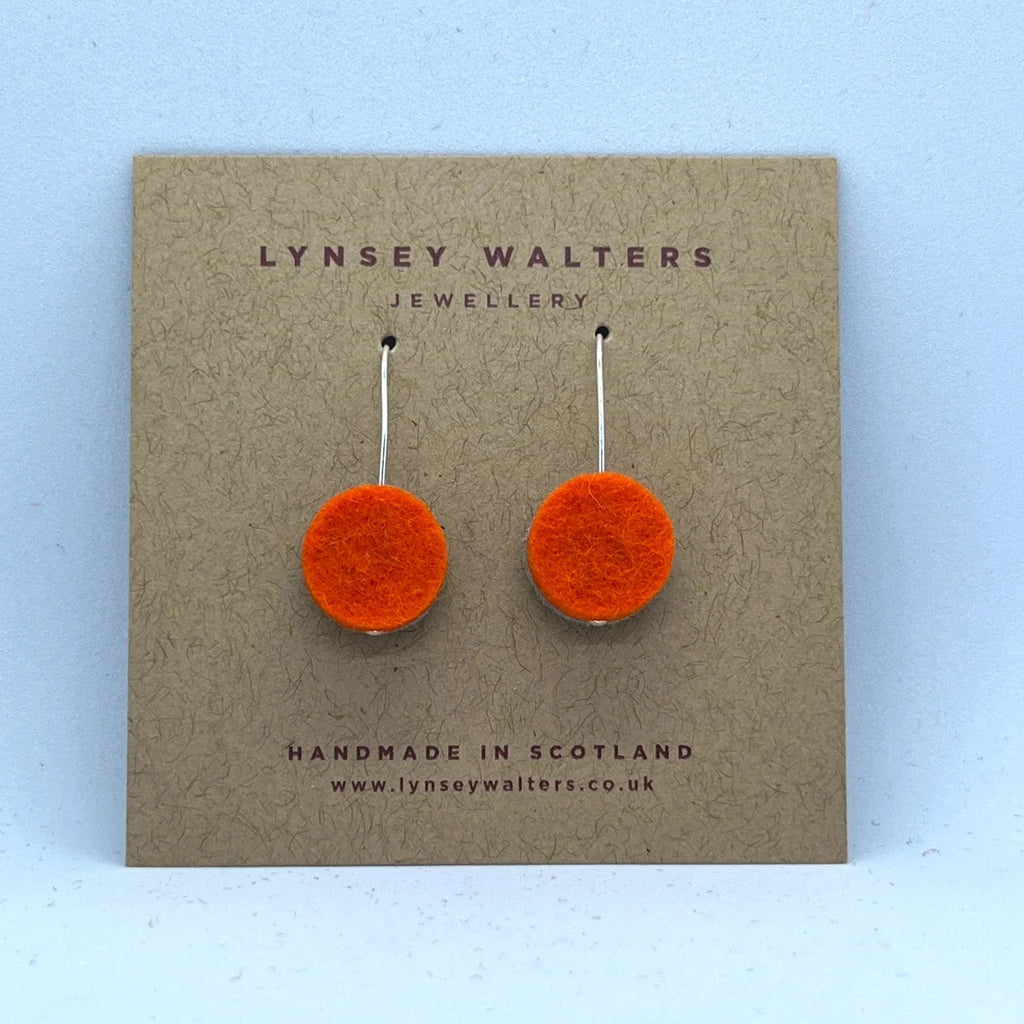 Pair of bright orange felt circle earrings with silver hooks, displayed on a brown card.