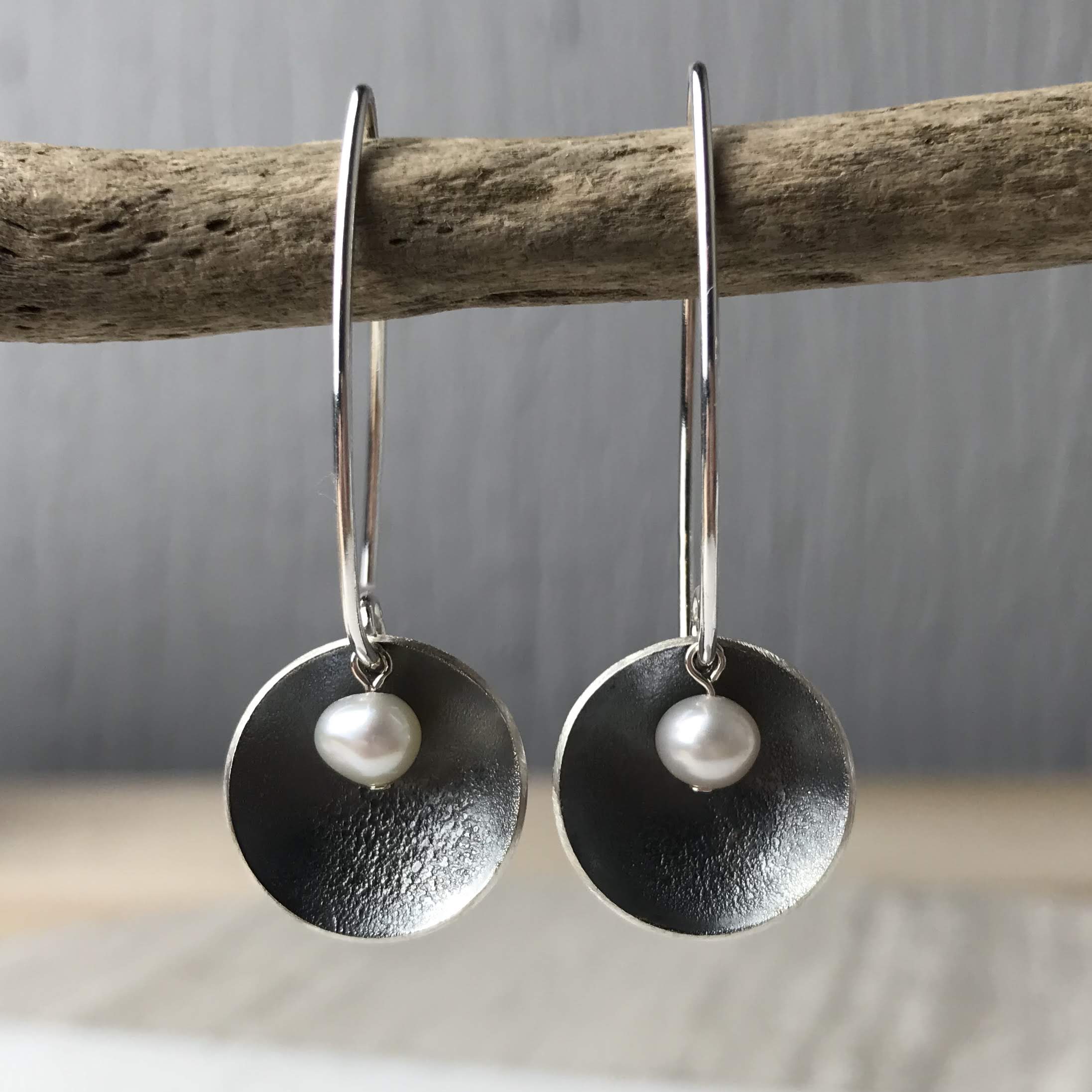 Pair of silver hoop earrings. Each hoop features a textured, dark-toned disc with a small pearl centered on it.