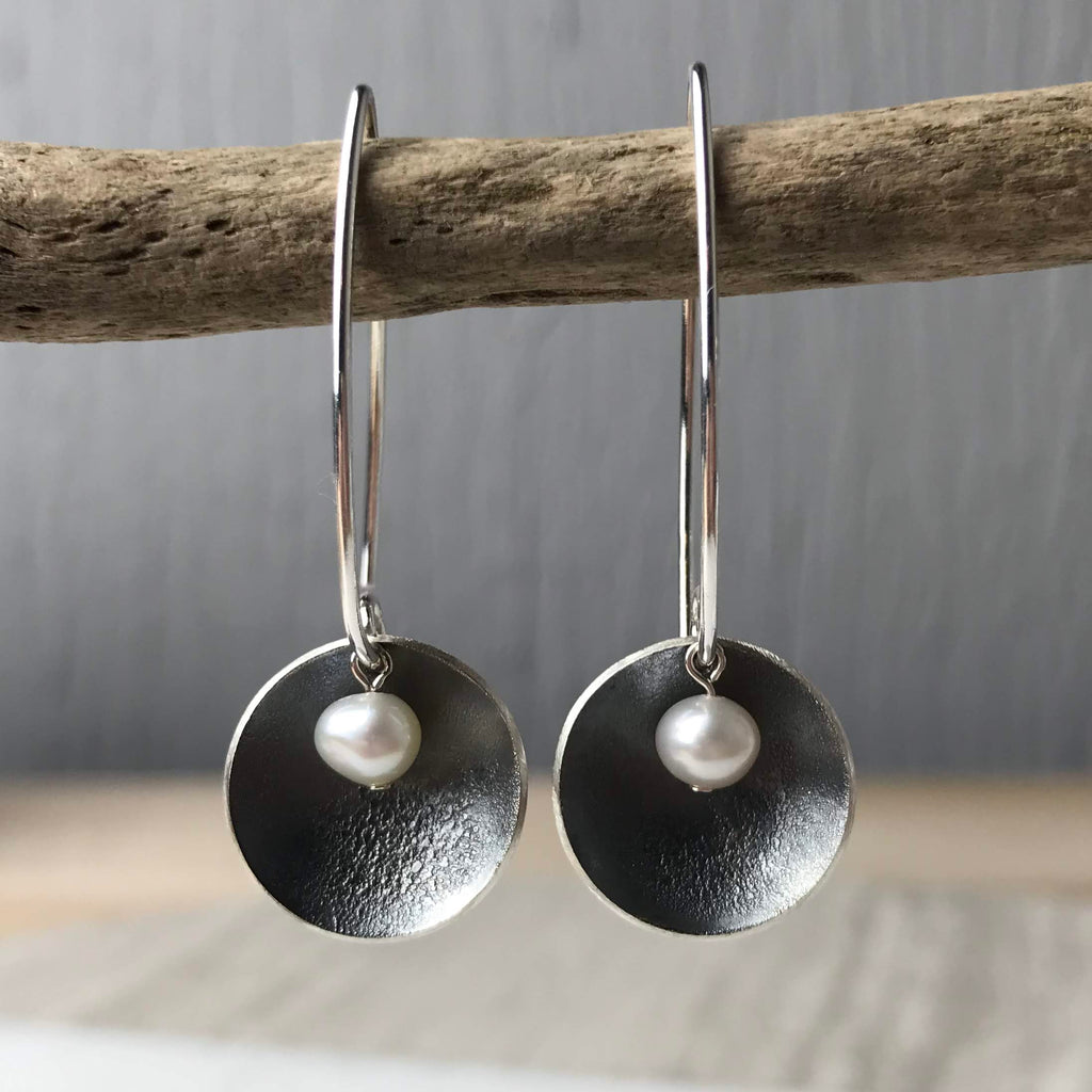 Pair of silver hoop earrings. Each hoop features a textured, dark-toned disc with a small pearl centered on it.