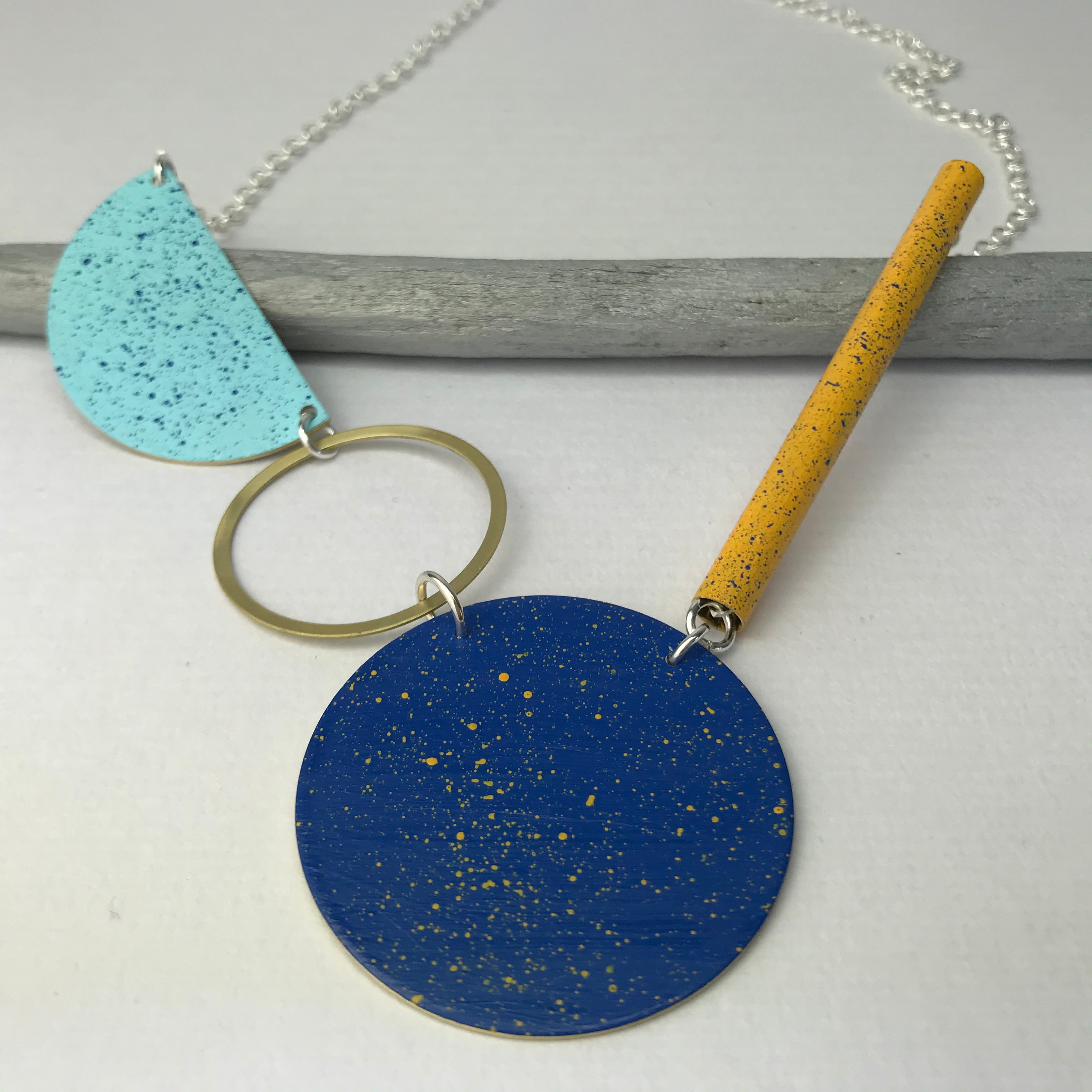 Modern statement necklace featuring blue, teal, and yellow speckled circular and cylindrical pendants on a silver chain.