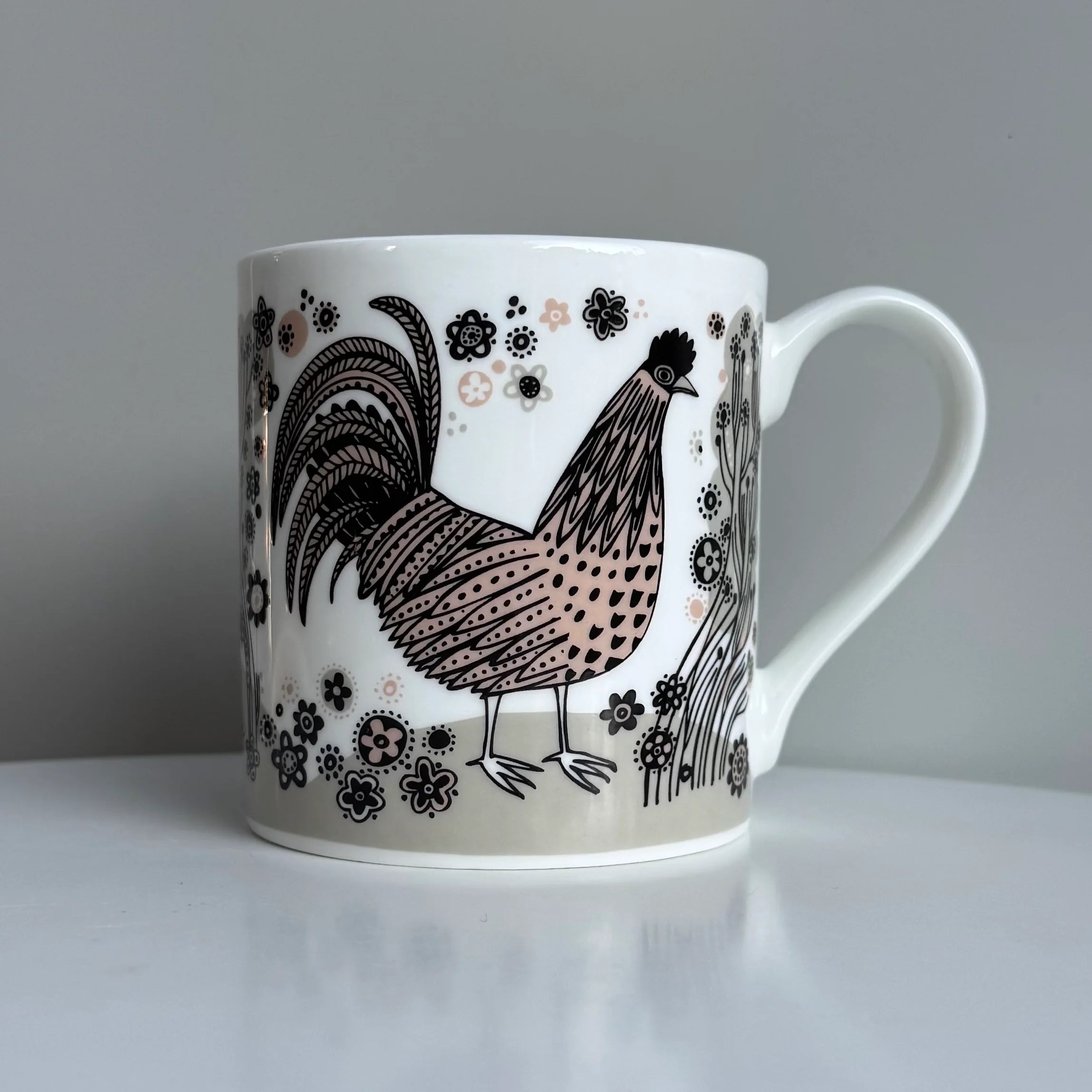 White mug with a black and brown rooster design on a white surface with a gray background